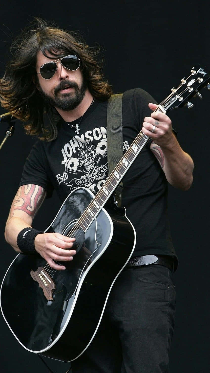 foo fighters 桌布 壁紙 — 一位長髮戴太陽眼鏡的音樂家在黑暗的舞台上熱情地彈奏黑色原聲結他。