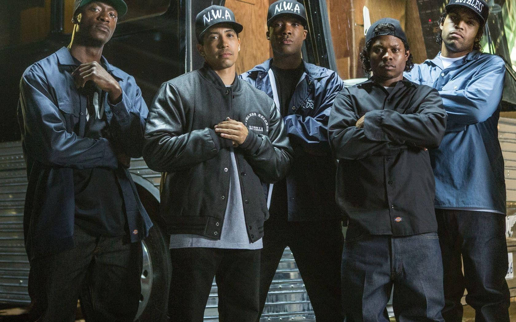 N.W.A. 2015年電影《Straight Outta Compton》 壁紙 — 五名戴著N.W.A帽子和穿著外套的男子在昏暗的車庫燈光下自信地站在一輛老爺車前。