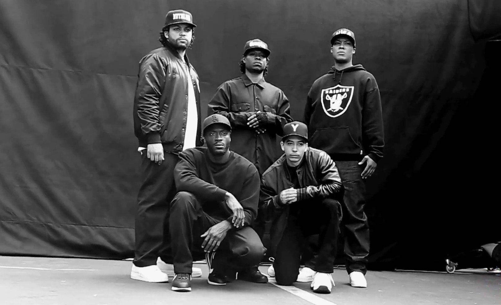N.W.A.《Straight Outta Compton》電影幕後花絮 壁紙 — 五個戴帽子和穿外套的男人在黑暗背景下自信地擺姿勢，拍攝成一張清晰的黑白工作室照片。