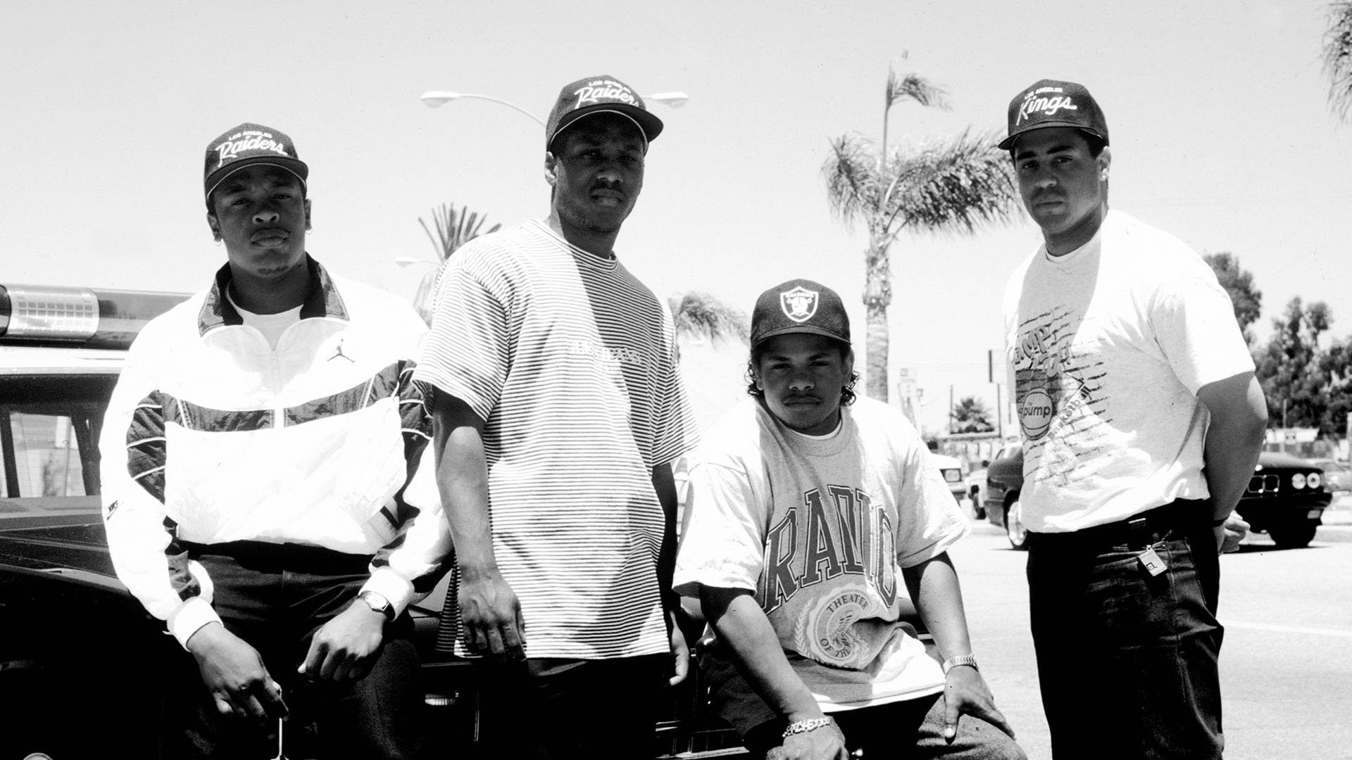 N.W.A.定義西岸嘻哈精神 壁紙 — 四個戴帽、身穿休閒服的男人在陽光明媚、有棕櫚樹的街道上自信地站在車旁擺姿勢。