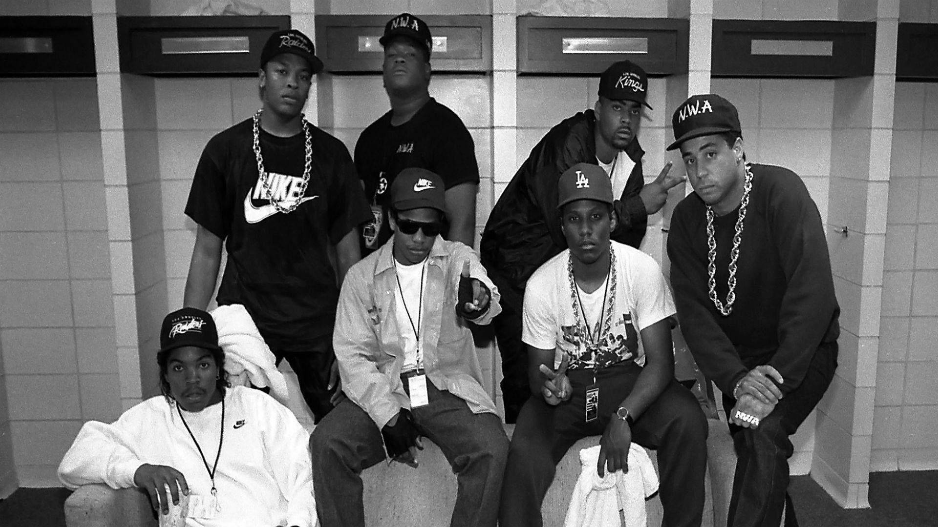 N.W.A.與The D.O.C.及Laylaw巡迴演出 壁紙 — 七名身穿90年代嘻哈服飾的男子在瓷磚更衣室中自信擺姿勢，佩戴大膽的鏈條和帽子。