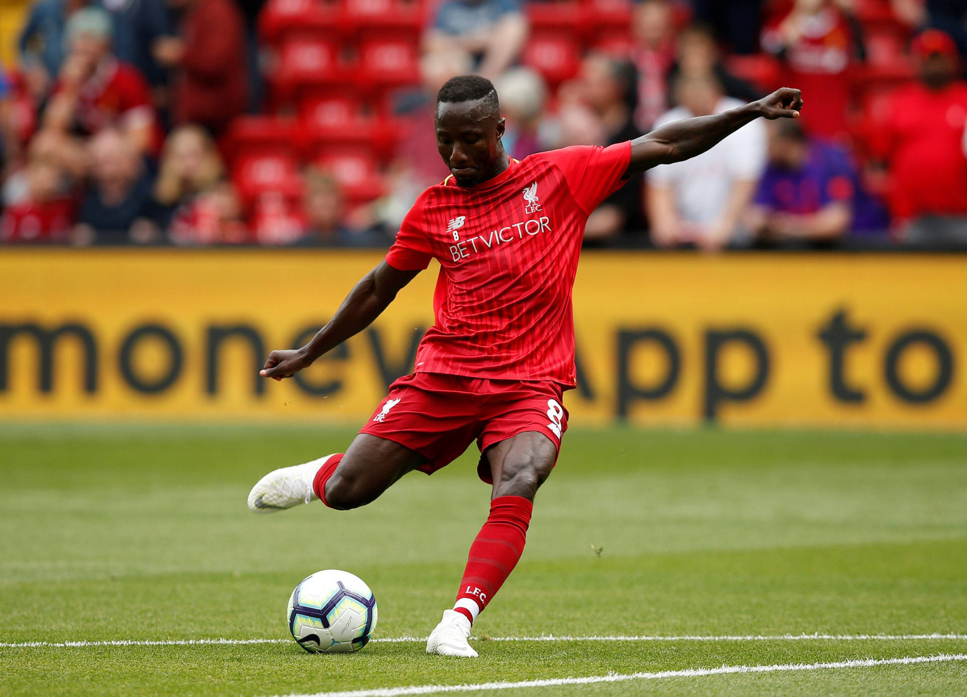 Naby Keita 強力射門 壁紙 — 一名身穿紅色球衣的利物浦足球員在充滿活力的綠色球場上踢足球，背景是模糊的紅色看台。