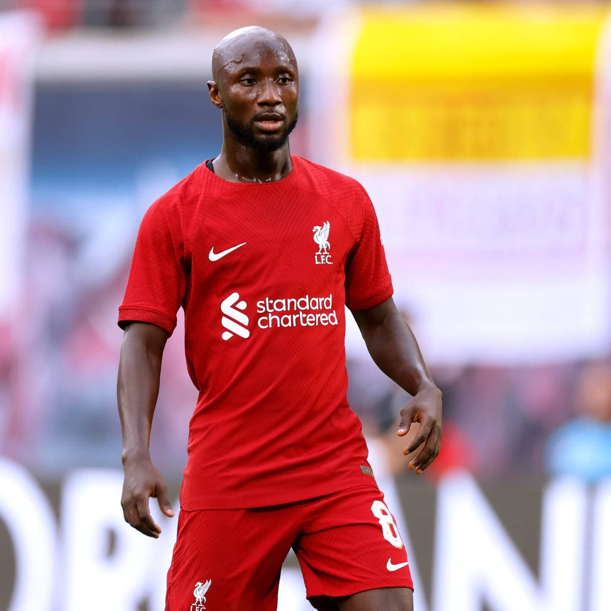 Naby Keita 穿紅色戰袍衝鋒 壁紙 — 一名專注的足球員身穿紅色利物浦FC球衣，站在陽光明媚的球場上，背景中的體育場看台模糊不清。