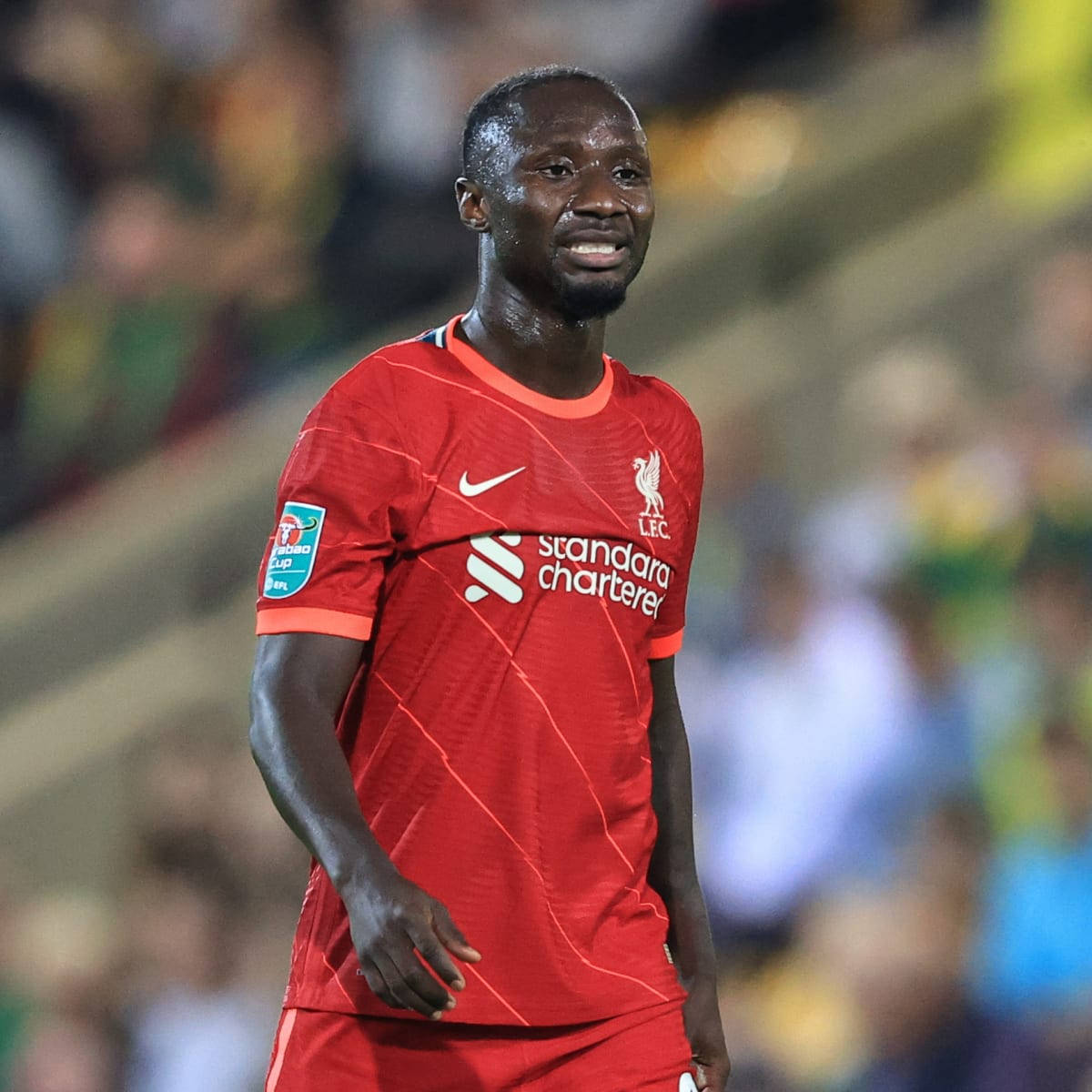 Naby Keita 紅色雙色球衣微笑 壁紙 — 一名身穿紅色球衣的利物浦足球員在夜間比賽中，在球場燈光下微笑，背景是模糊的人群。