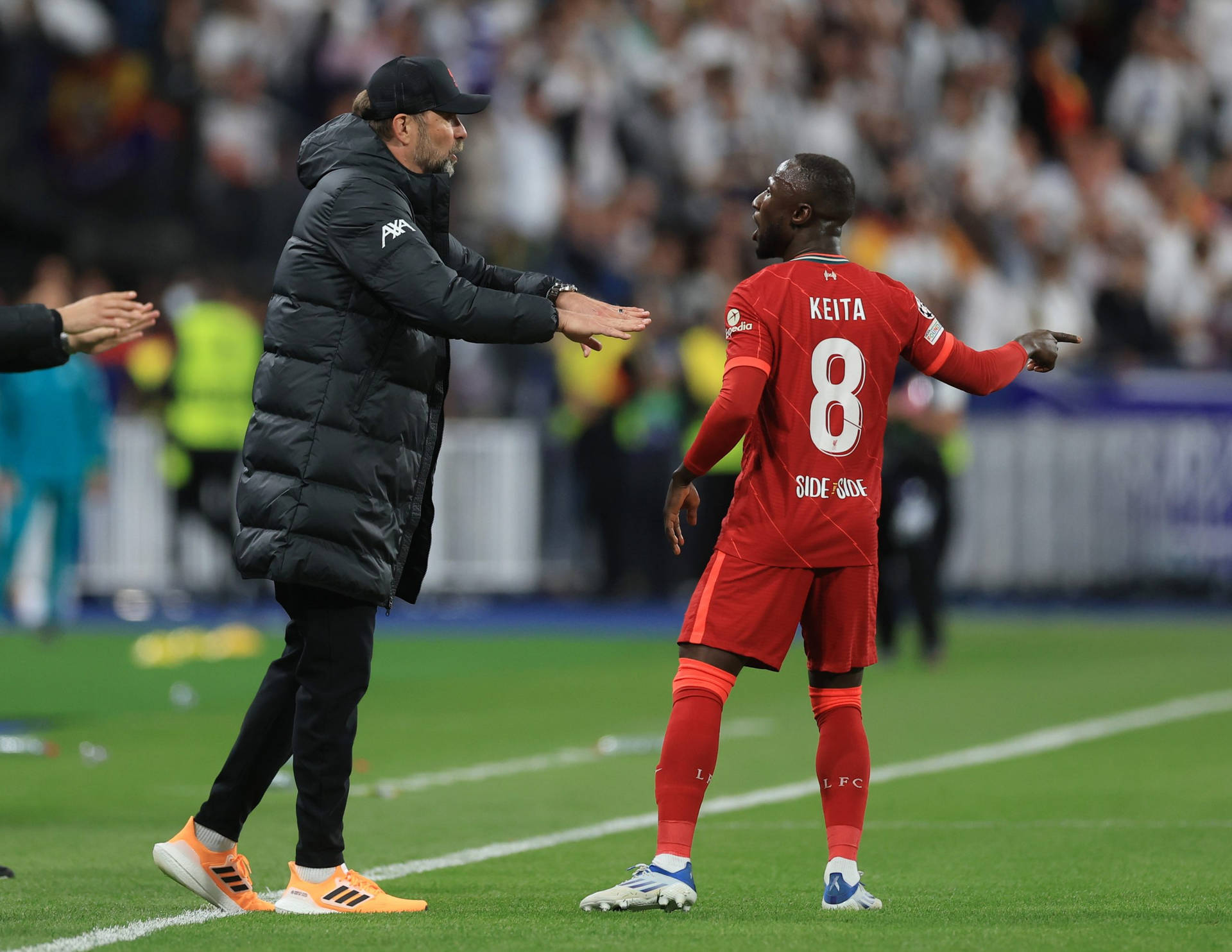 Naby Keita 與裁判交談 壁紙 — 一位身穿黑色羽絨外套的足球教練，向一位身穿紅色球衣、名叫基達的球員做手勢，背景是綠色球場和模糊的體育場人群。