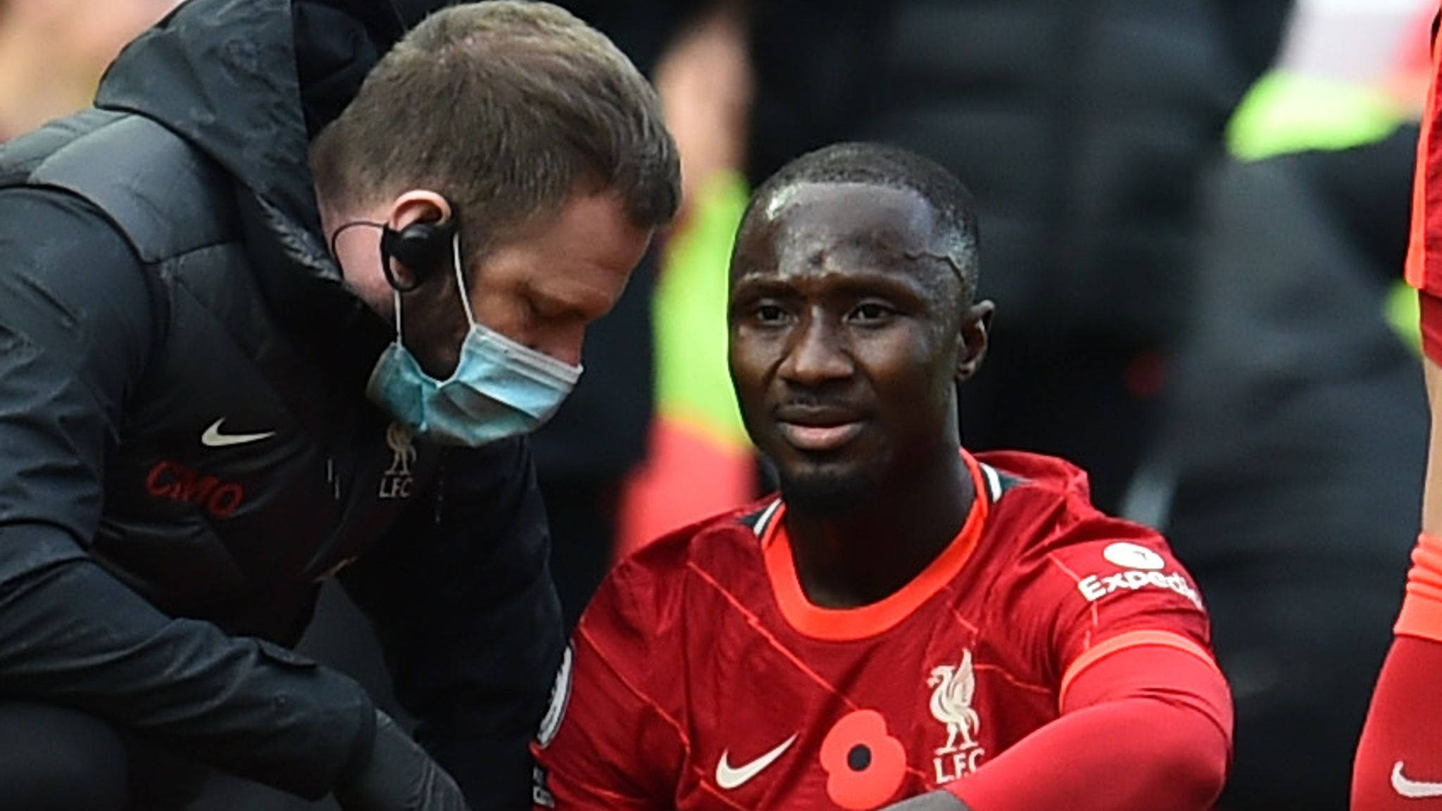 Naby Keita 被安慰 壁紙 — 一名身穿紅色球衣的利物浦足球員坐在邊線，一名戴口罩的醫護人員俯身檢查他的情況。