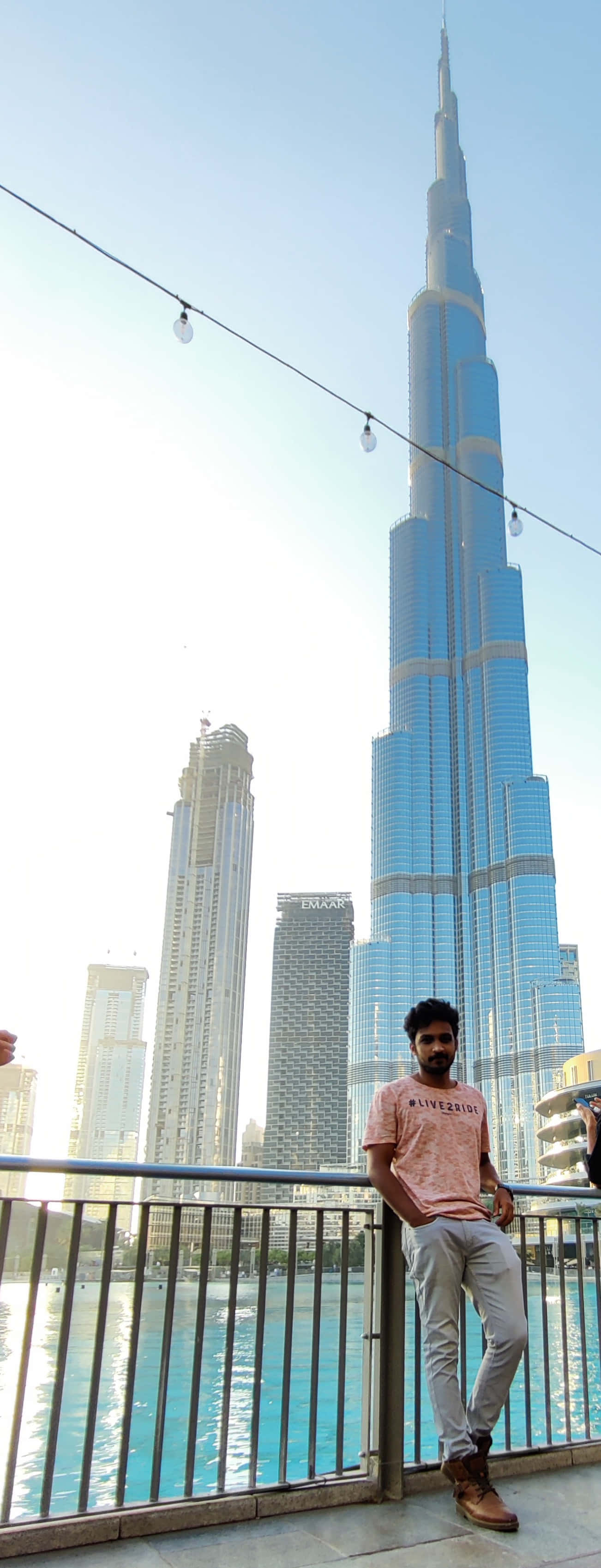 Burj Khalifa 桌布 壁紙 — 一名男子隨意靠在欄杆上，俯瞰水面，高聳的哈利法塔主宰了明亮的白天天際線。