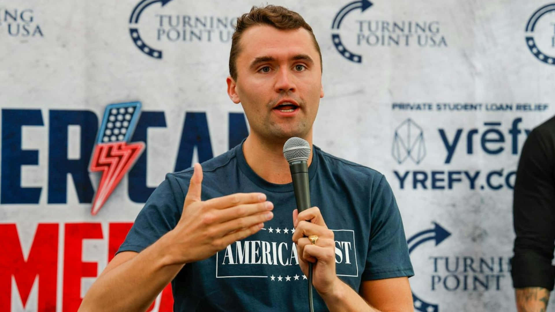  壁紙 — 一名身穿藍色「AMERICANIST」T恤的男子在Turning Point USA活動中對著麥克風熱情發言。
