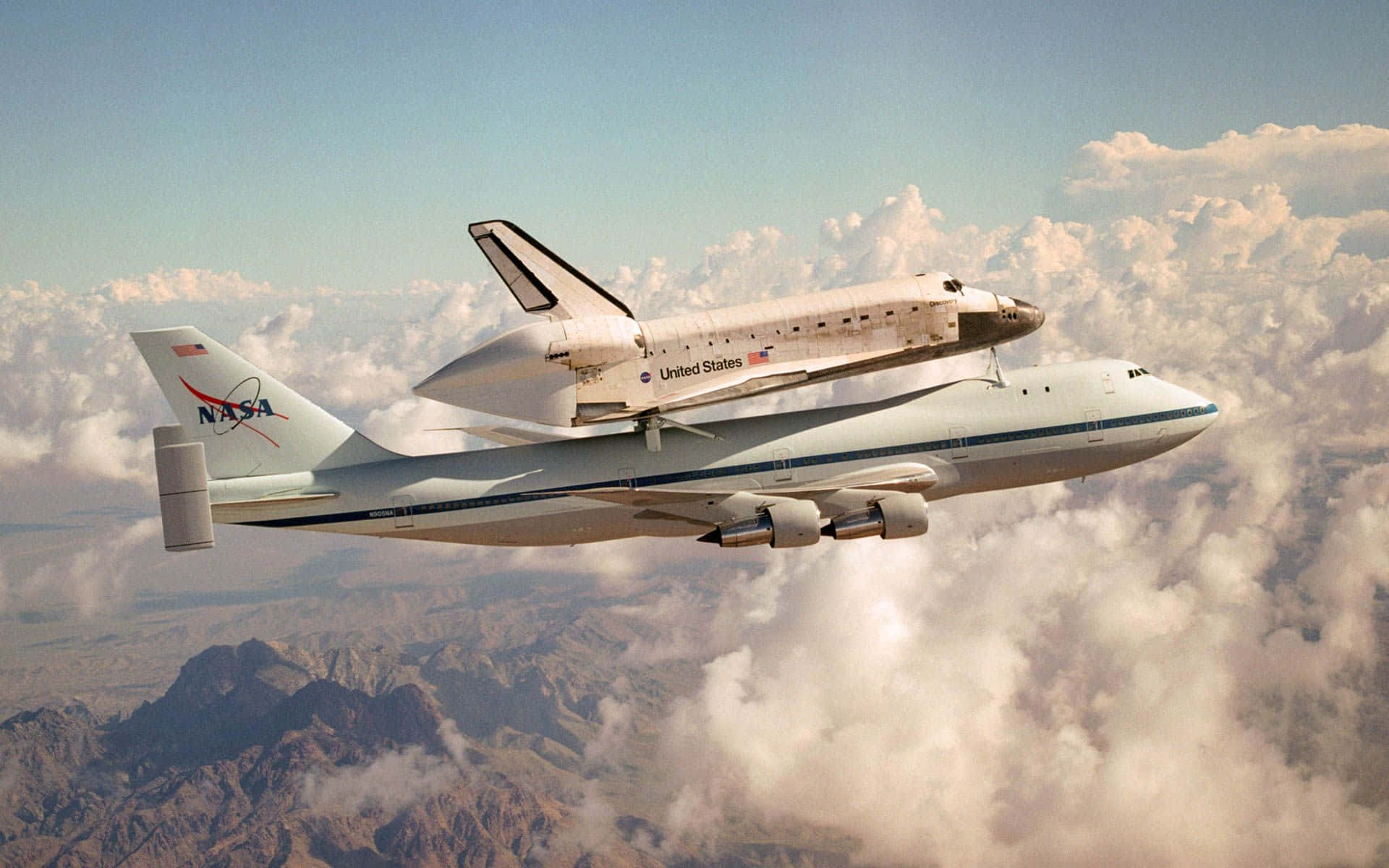太空梭與NASA運輸機翱翔天際 壁紙 — 一架NASA太空穿梭機安裝在改裝的波音747上，在蓬鬆的雲層上方翱翔，下方是崎嶇的山脈。