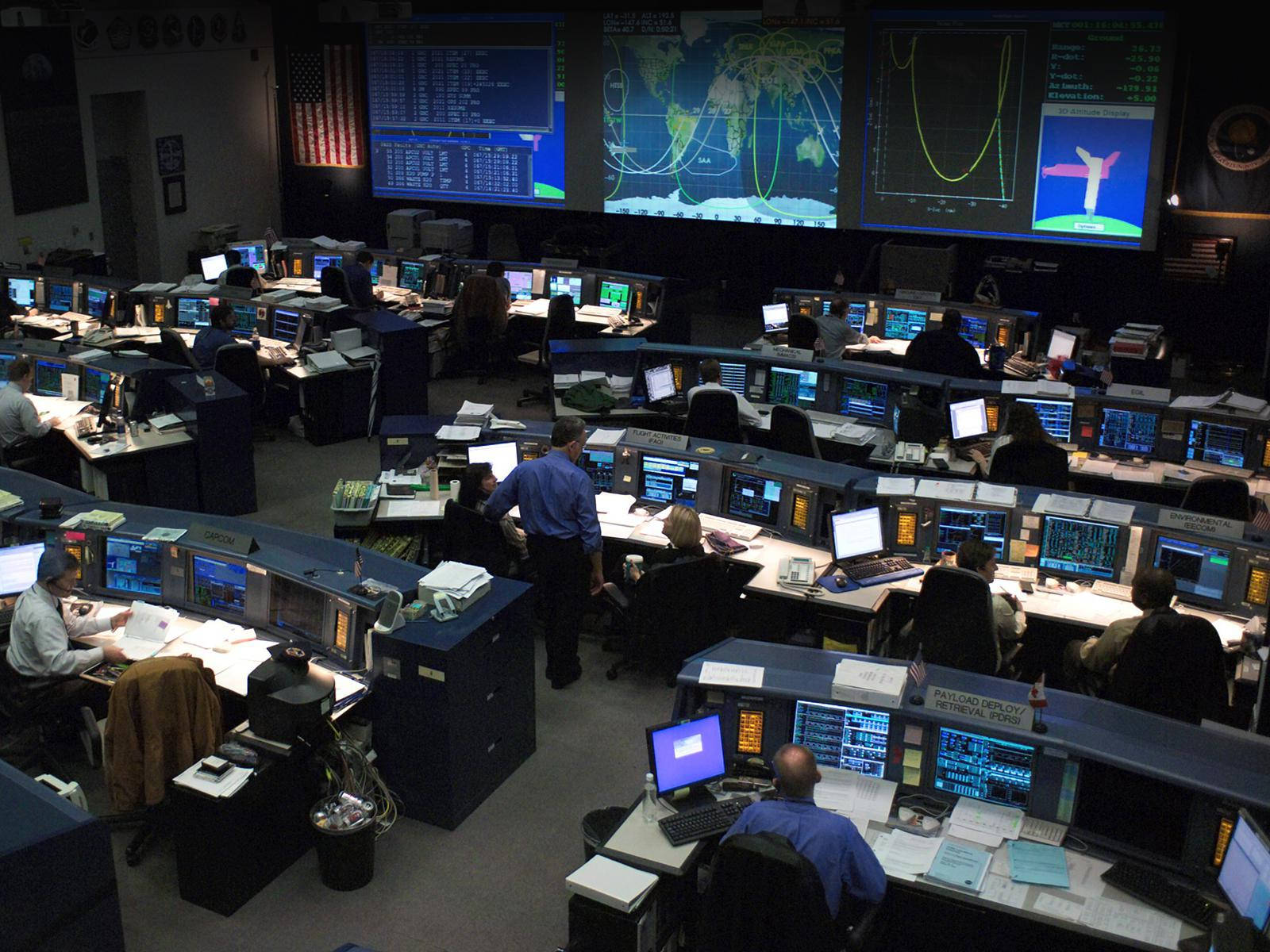 NASA休士頓電腦 壁紙 — 燈光昏暗的NASA任務控制室，工程師們在電腦工作站前監控大屏幕上的全球數據。