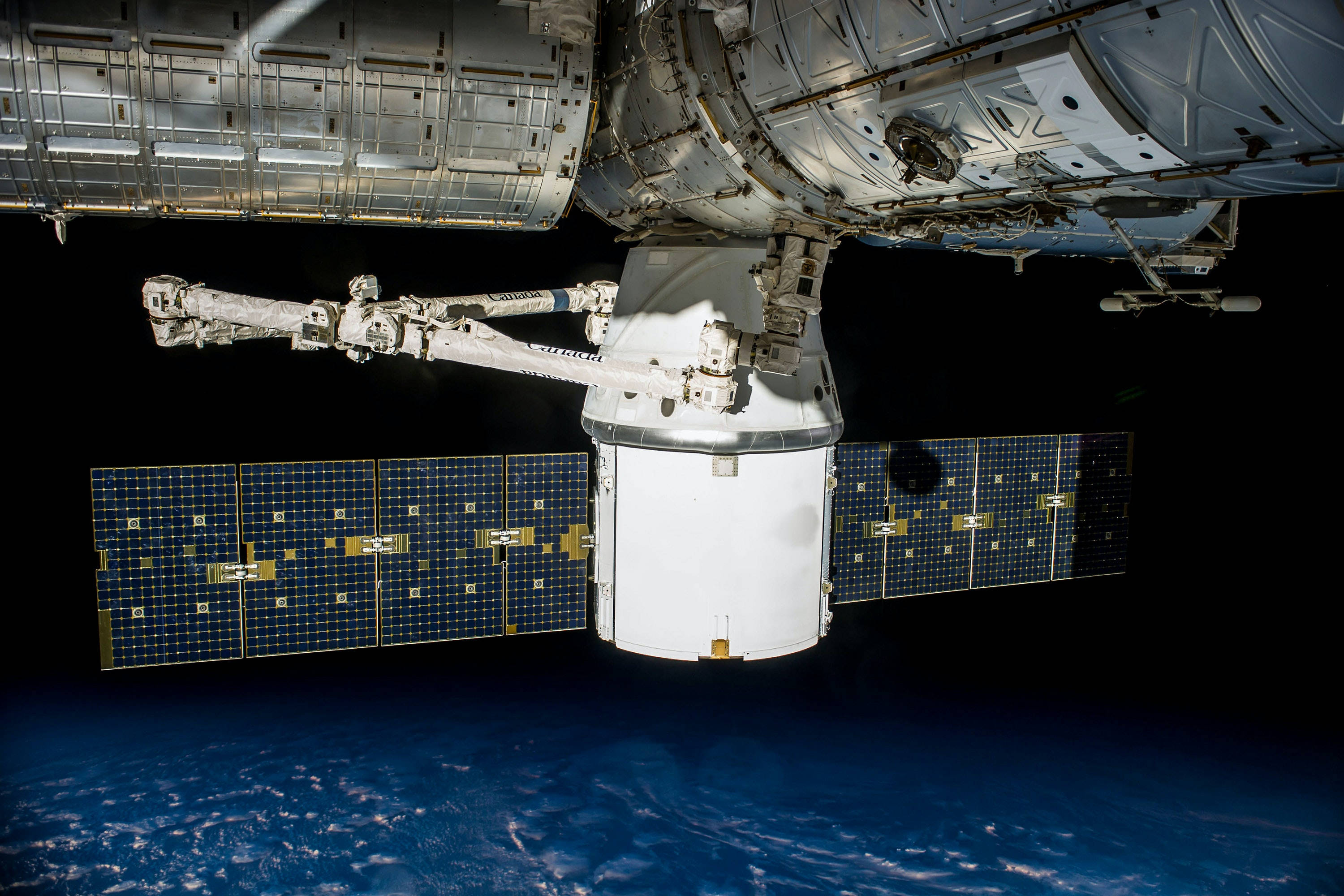 NASA休斯頓衛星 壁紙 — 一艘帶有太陽能板的太空船從太空站模組延伸出來，面對地球藍色彎曲地平線之上的黑色虛空。