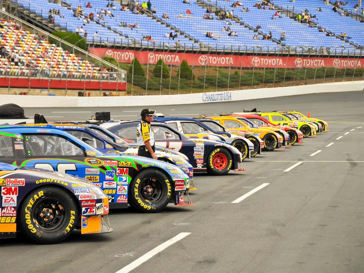 NASCAR賽車停泊 壁紙 — 一排色彩鮮豔的NASCAR賽車整齊排列在賽道上，旁邊站著一名賽道工作人員，天空部分多雲。