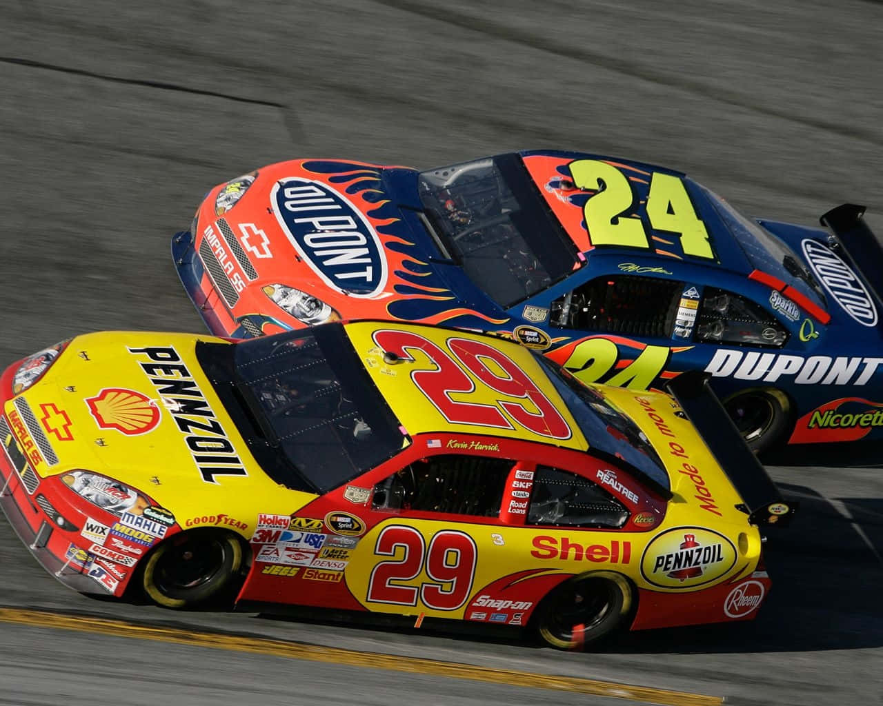 NASCAR賽車貼身跟隨 壁紙 — 兩輛帶有鮮豔贊助商貼紙的NASCAR賽車在明亮的日光下並排疾馳在賽道上。