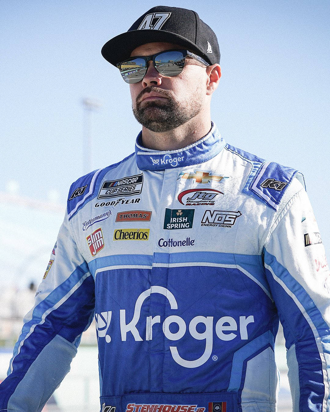 專業NASCAR車手瑞奇·斯滕豪斯小將肖像 壁紙 — 一位身穿藍白 Kroger 賽車服的 NASCAR 車手，戴著太陽眼鏡和帽子，在明亮的日光下自信地站立。