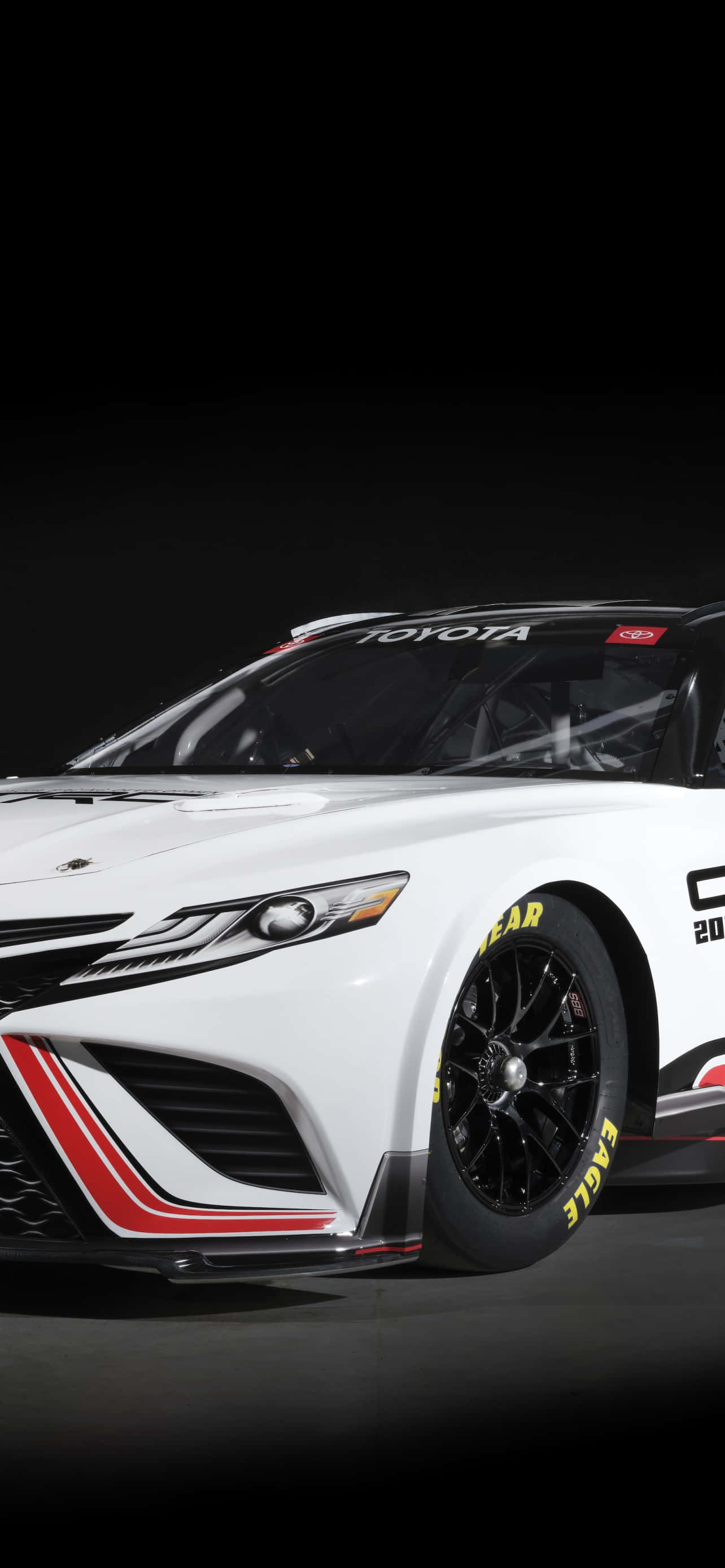 在你的Nascar iPhone上衝向終點線 壁紙 — 一架流線型白色豐田賽車，帶有紅色和黑色點綴以及黃色Eagle輪胎，在深色工作室背景中展示。