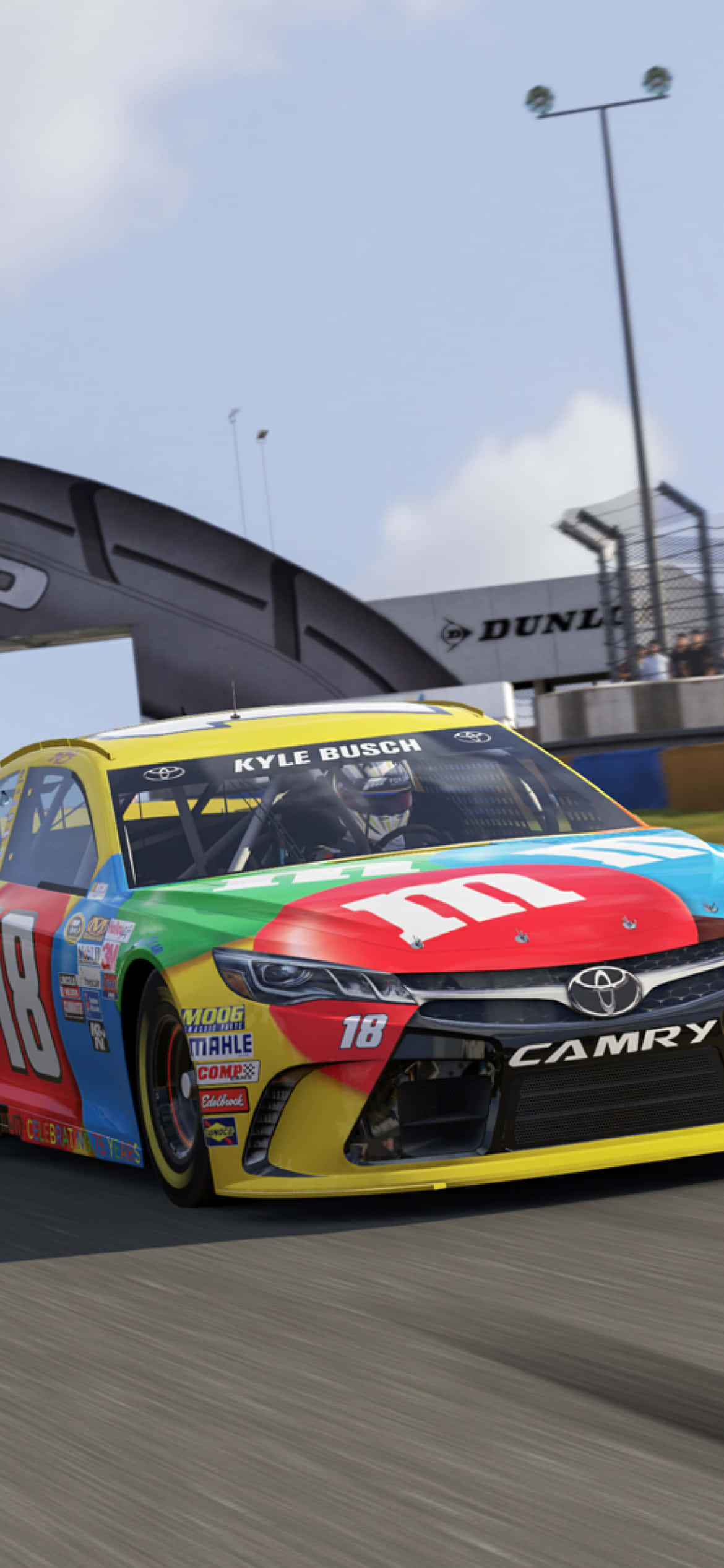 圖片 展示你的NASCAR精神，配上Nascar iPhone 壁紙 — 一輛色彩繽紛的M&M's品牌NASCAR賽車在晴朗的天空下，由Kyle Busch駕駛在賽道上飛馳。