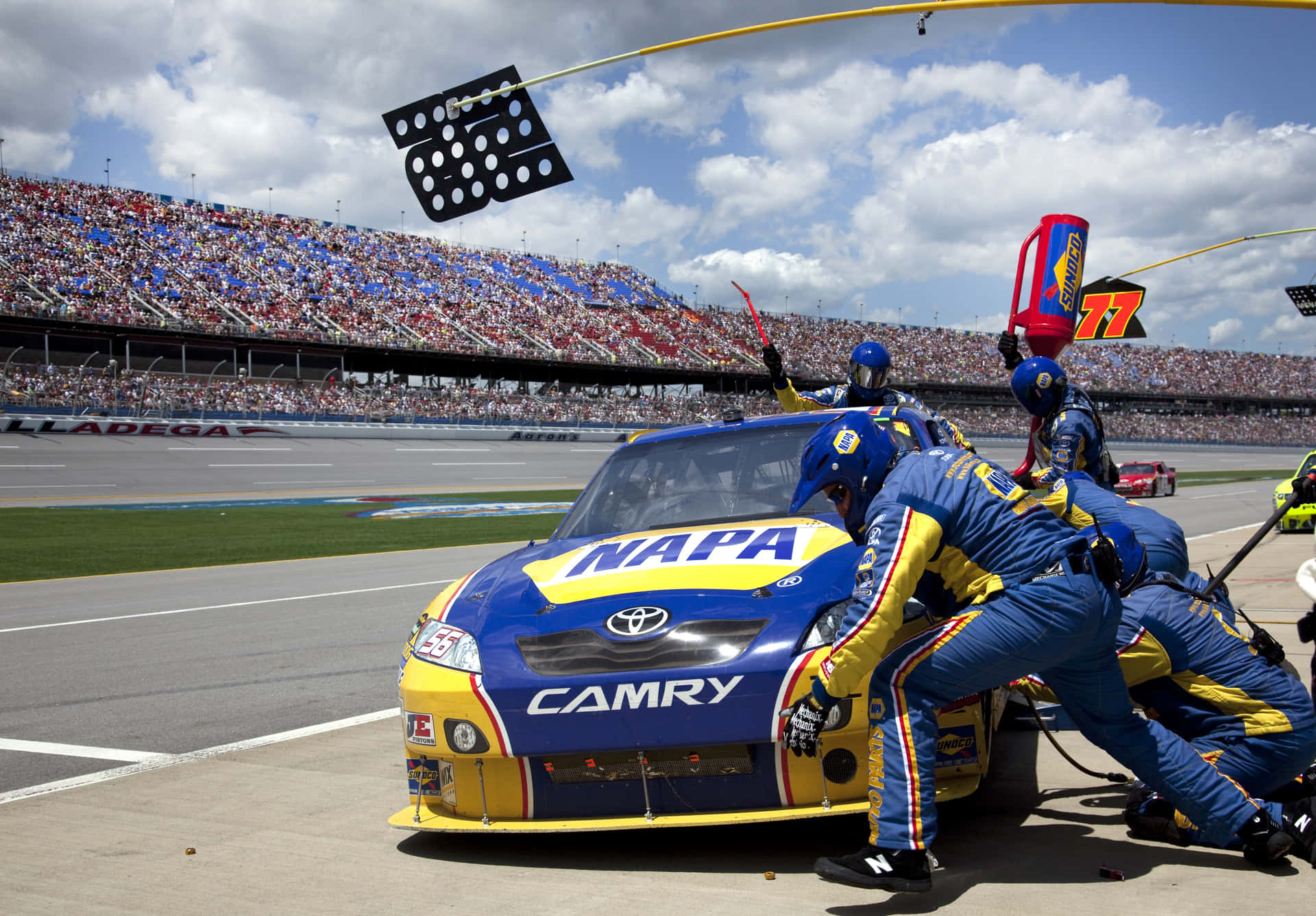 NASCAR賽道上的驚險賽事 壁紙 — 一架藍黃色的NASCAR豐田Camry賽車正在維修站停靠，身穿藍黃色制服的工作人員在多雲的天空下，於擠滿觀眾的賽道上更換輪胎。