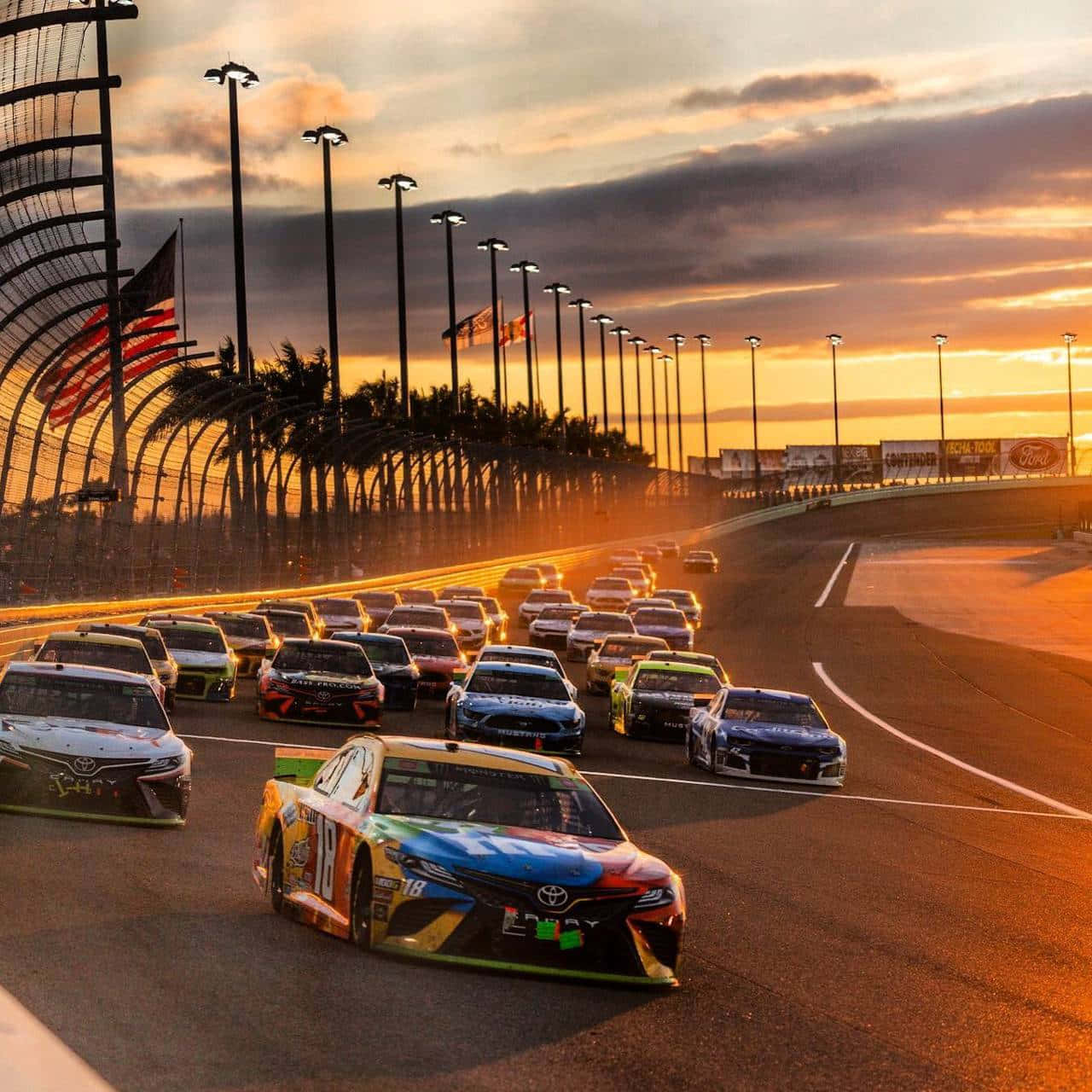 NASCAR賽車在日落時分的賽道上飛馳，背景有棕櫚樹和旗幟。