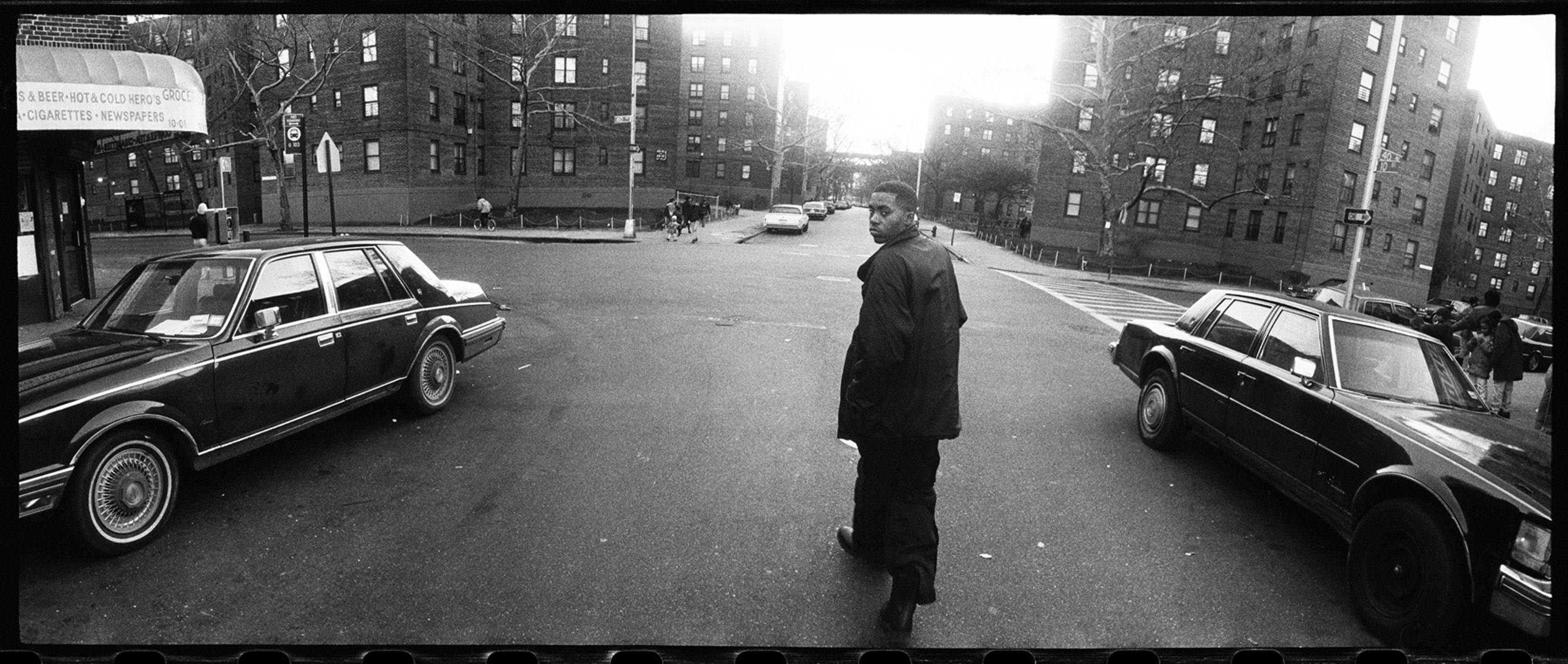 Nas《Time Is Illmatic》專輯海報 壁紙 — 一名男子在陽光普照的都市街道上行走，兩旁是經典汽車和磚造公寓大樓，以黑白呈現。