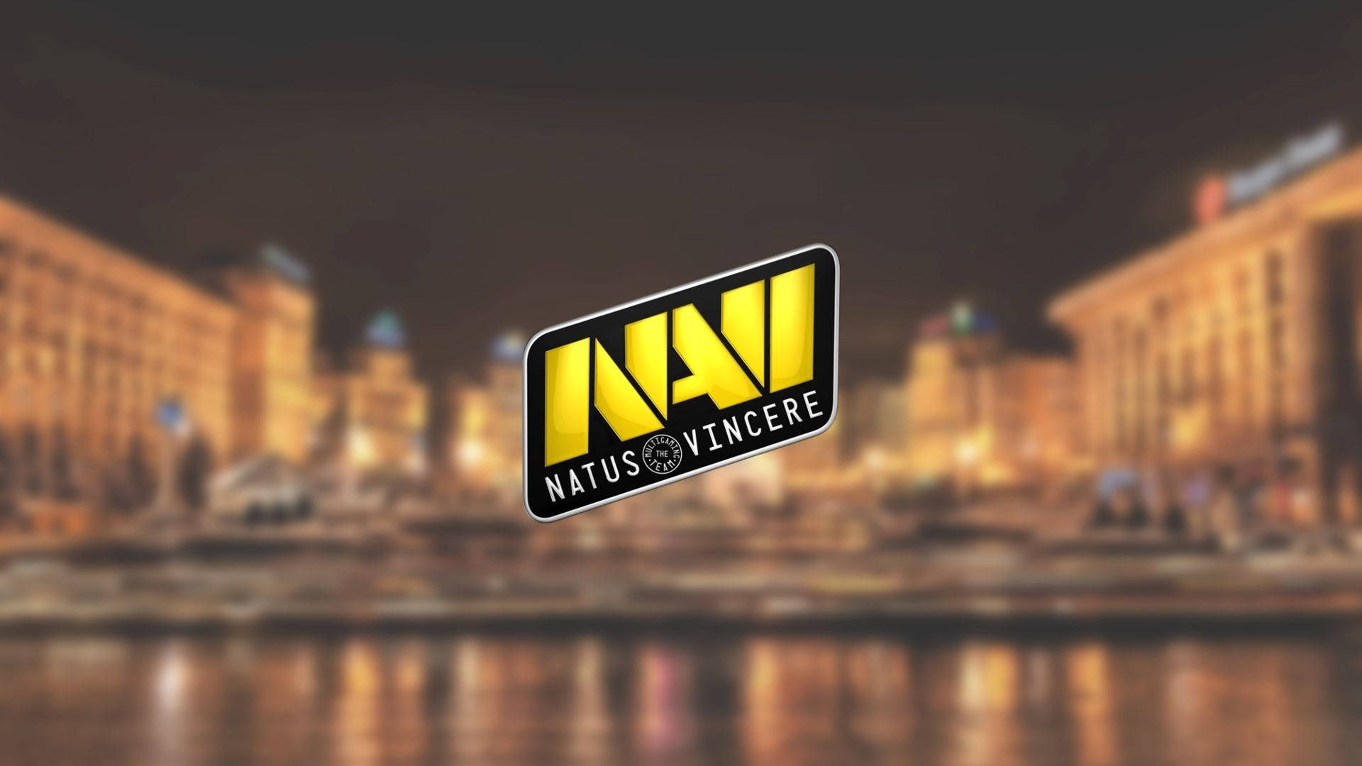 Natus Vincere 城市夜景 壁紙 — Natus Vincere 標誌在模糊的夜間城市景觀和水面上的溫暖倒影中，以鮮明的黃色發光。