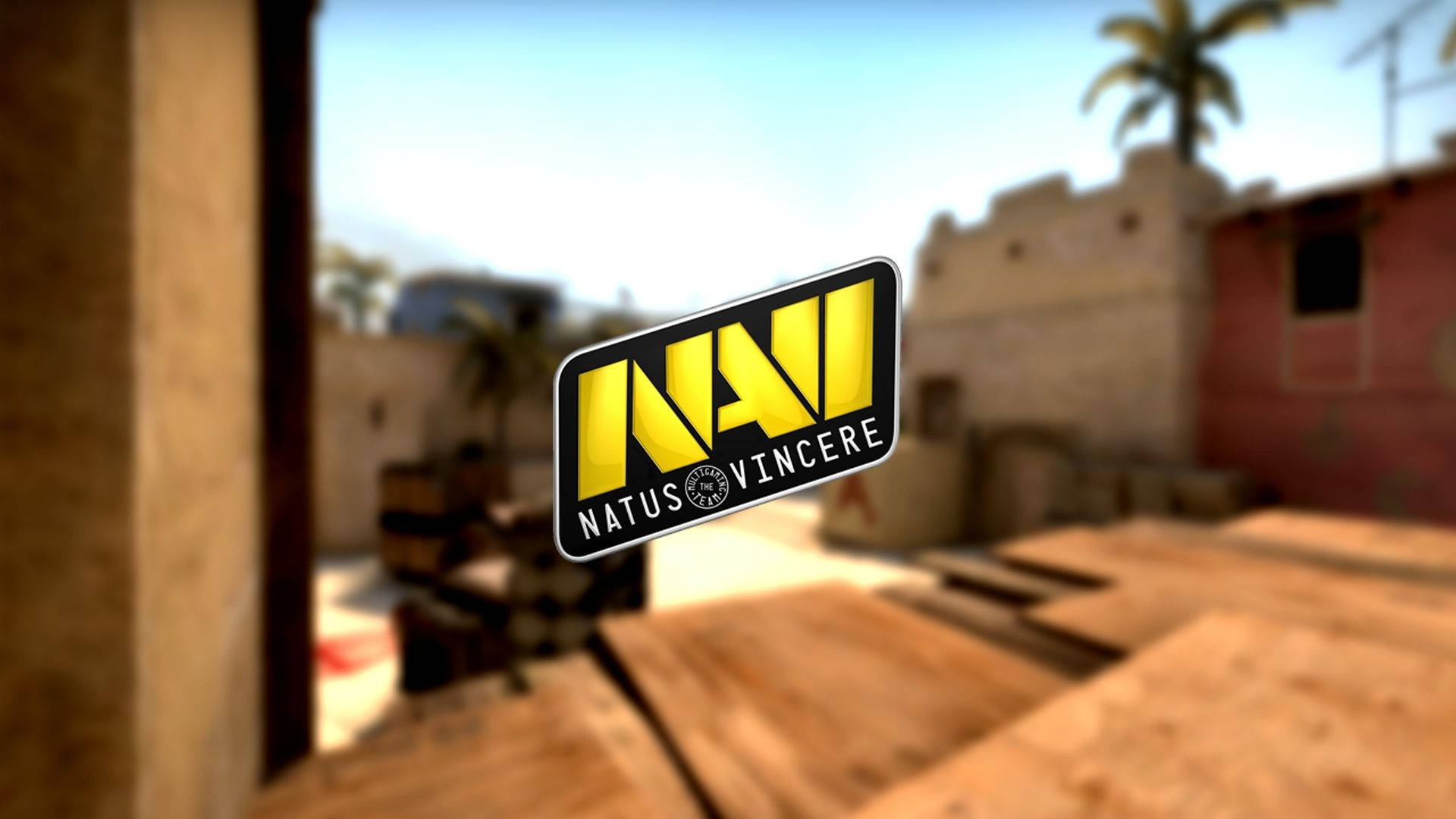 Natus Vincere 战場之上 壁紙 — 一個鮮黃色的Natus Vincere標誌漂浮在模糊的沙漠主題遊戲地圖前，地圖上有木板和石頭建築。