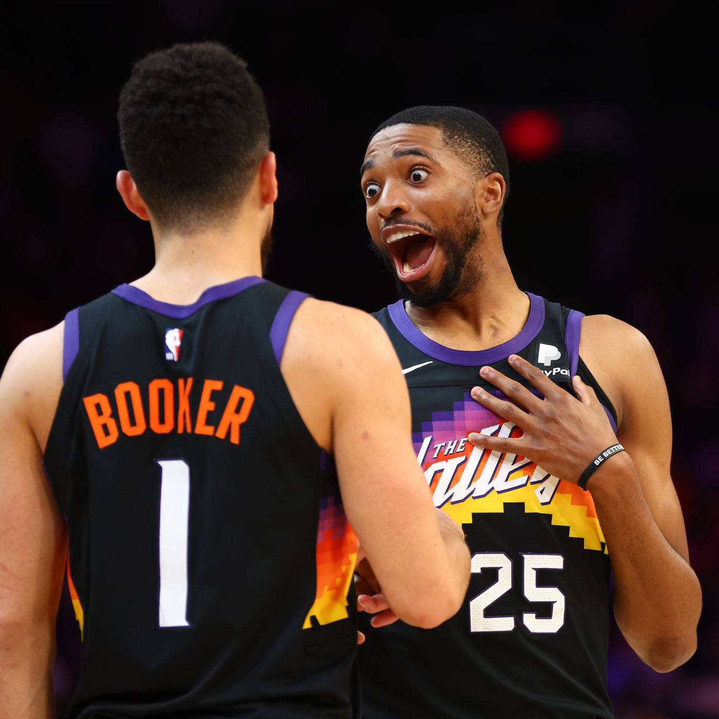 NBA 25號 米卡爾·布里奇斯 壁紙 — 兩名身穿鳳凰城太陽隊球衣的籃球運動員在場館燈光下的比賽中表現出震驚和興奮。