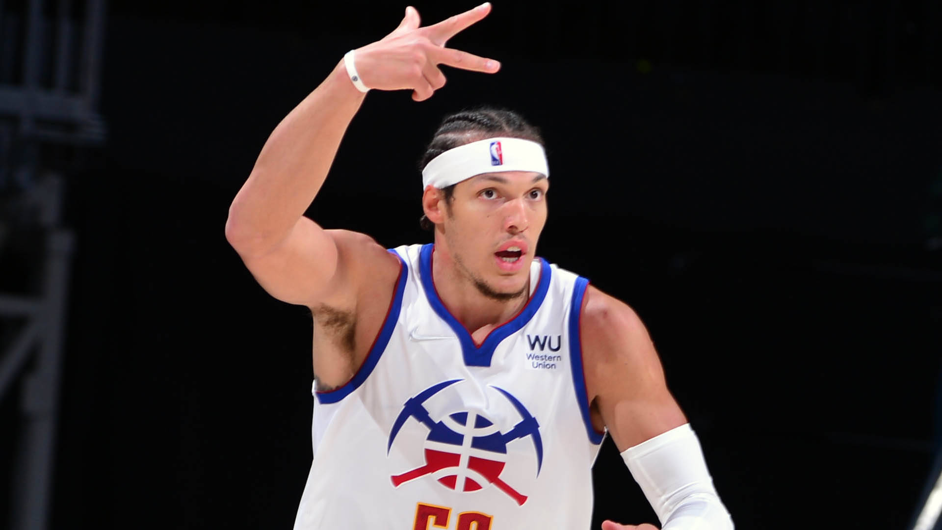 NBA球員阿隆·戈登 壁紙 — 一名身穿白色球衣帶有藍色和紅色點綴的籃球運動員在明亮的場館燈光下比賽中做出和平手勢。