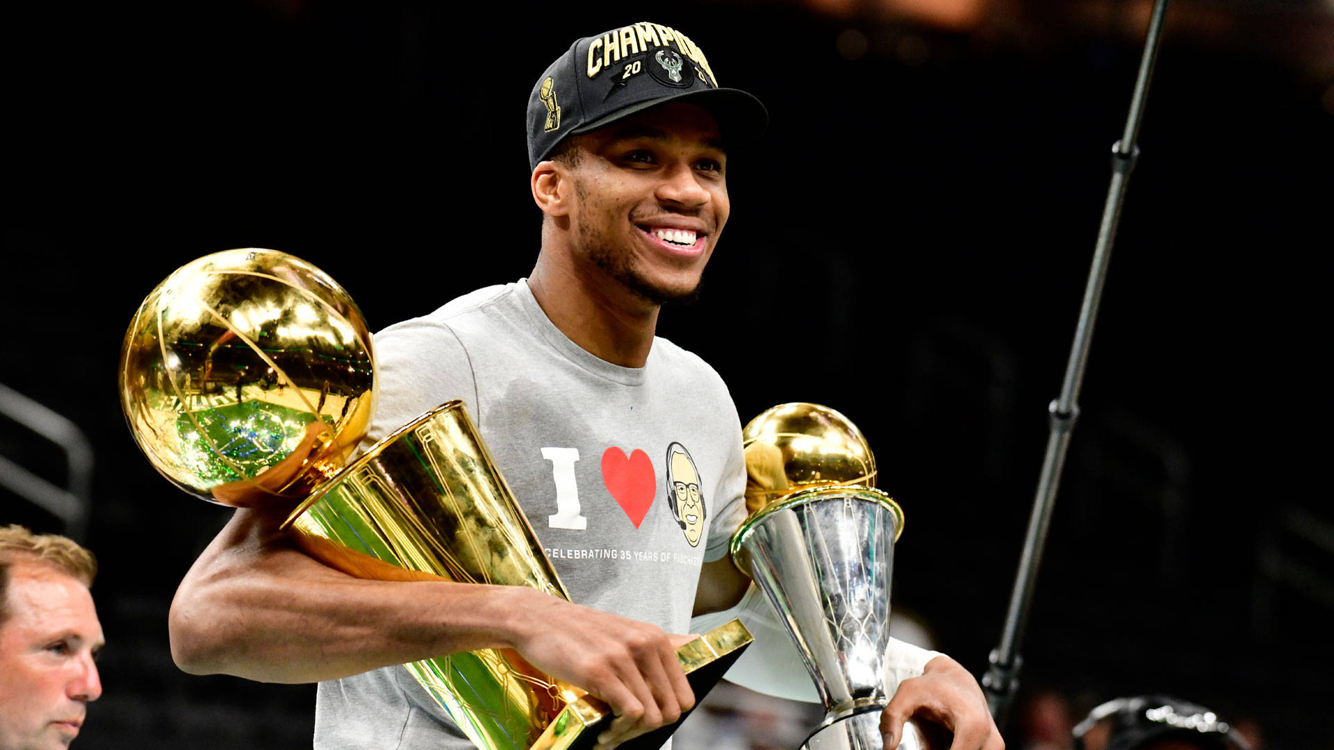 NBA總決賽吉安尼斯捧獎 壁紙 — 一位勝利的籃球運動員在場館燈光下，手持兩個閃閃發光的冠軍獎盃，笑容滿面。