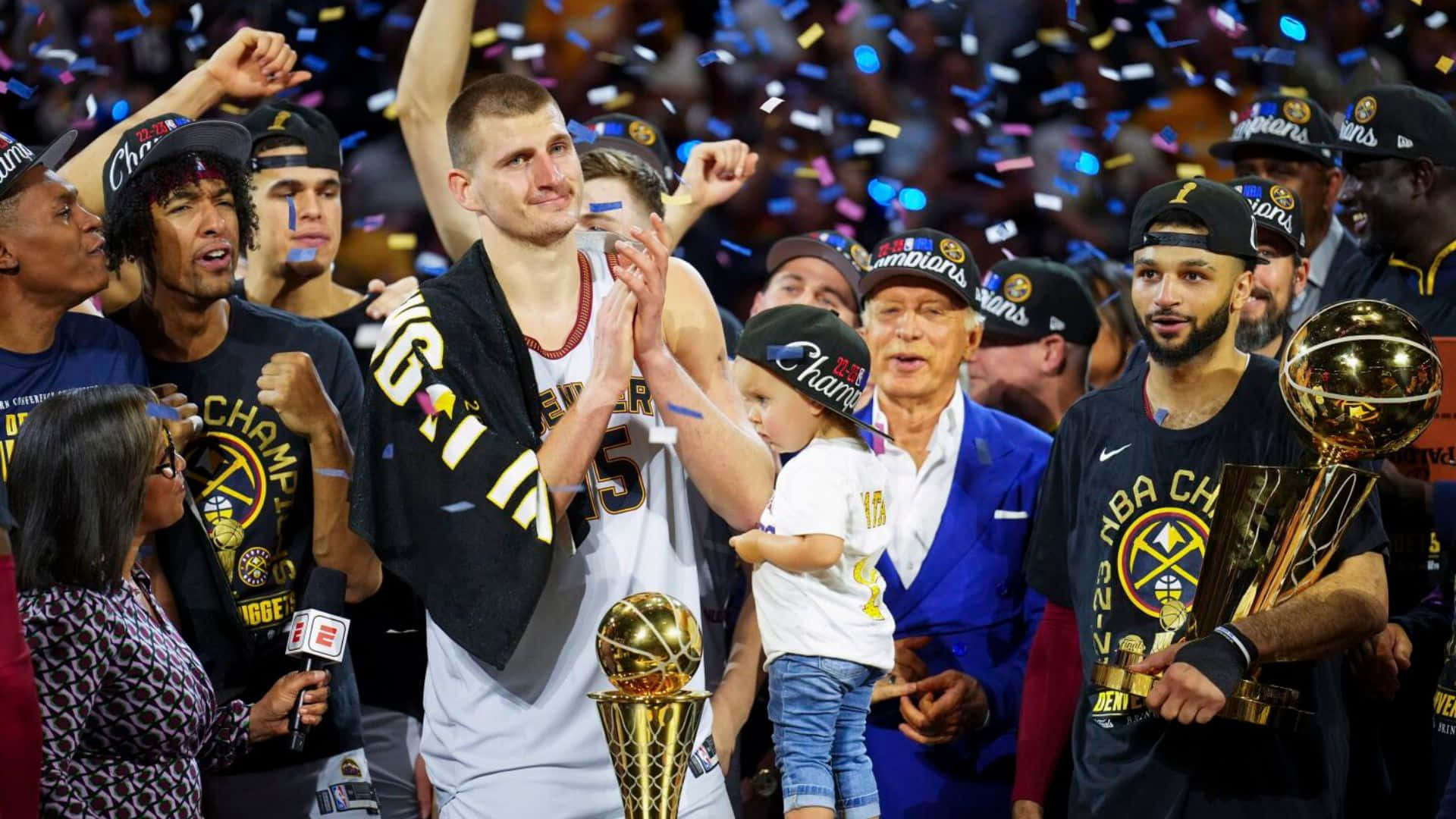 NBA Champions 桌布 壁紙 — NBA冠軍以獎盃和五彩紙屑慶祝，一名戴著'Champions'帽子的孩子站在球員之間。