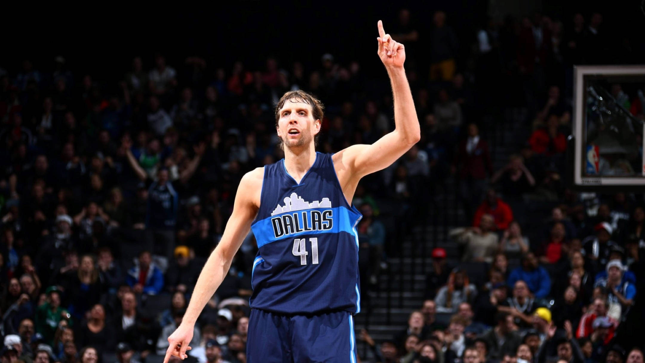 NBA德克·諾維茨基 壁紙 — 一名身穿達拉斯41號球衣的籃球運動員在明亮的場館燈光下比賽時，充滿強度地向上指。