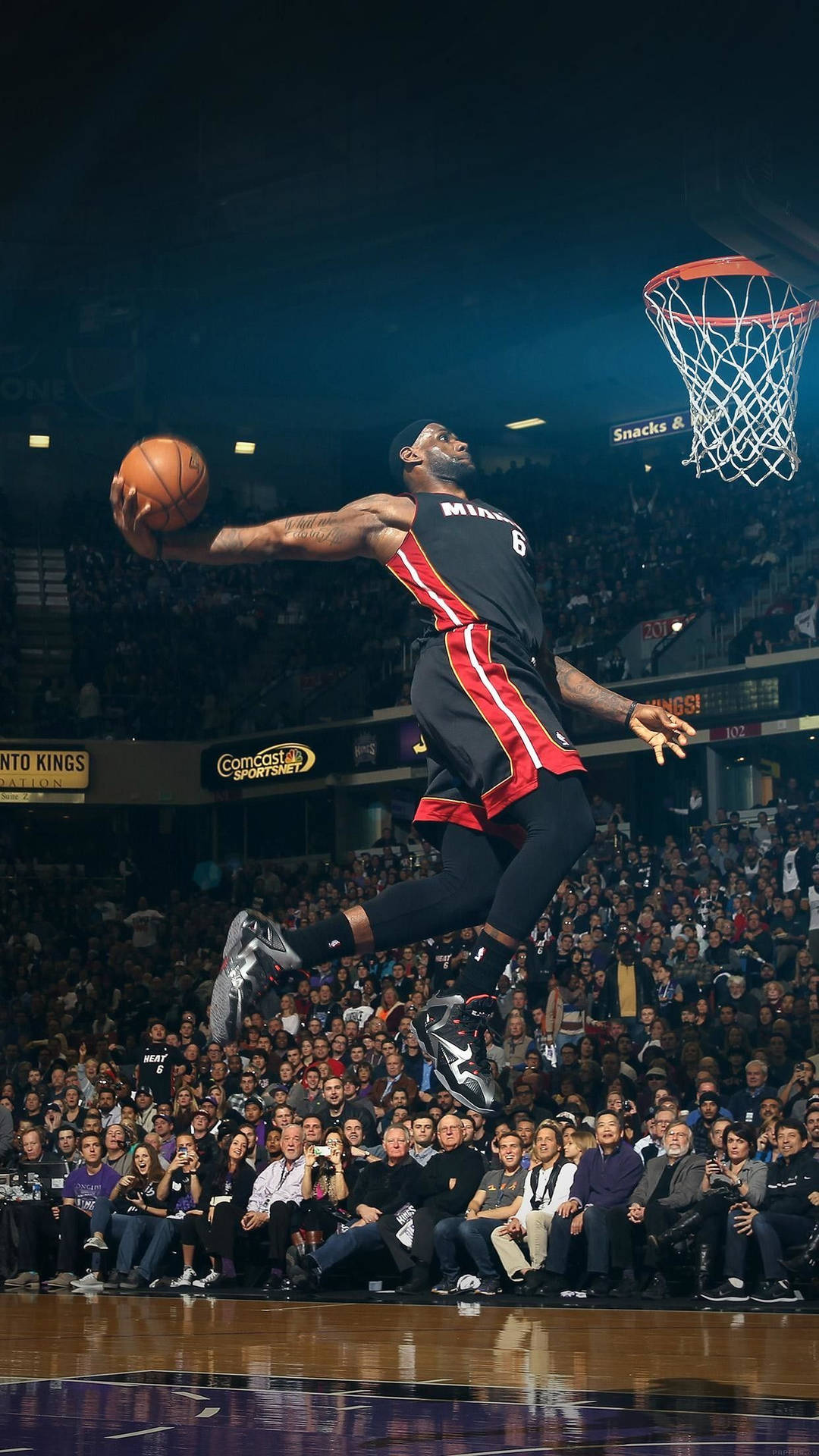 NBA iPhone 勒布朗·詹姆斯灌籃 壁紙 — 一名身穿黑紅色球衣的籃球運動員在座無虛席的球場比賽中，手持籃球騰空而起，飛向籃框。