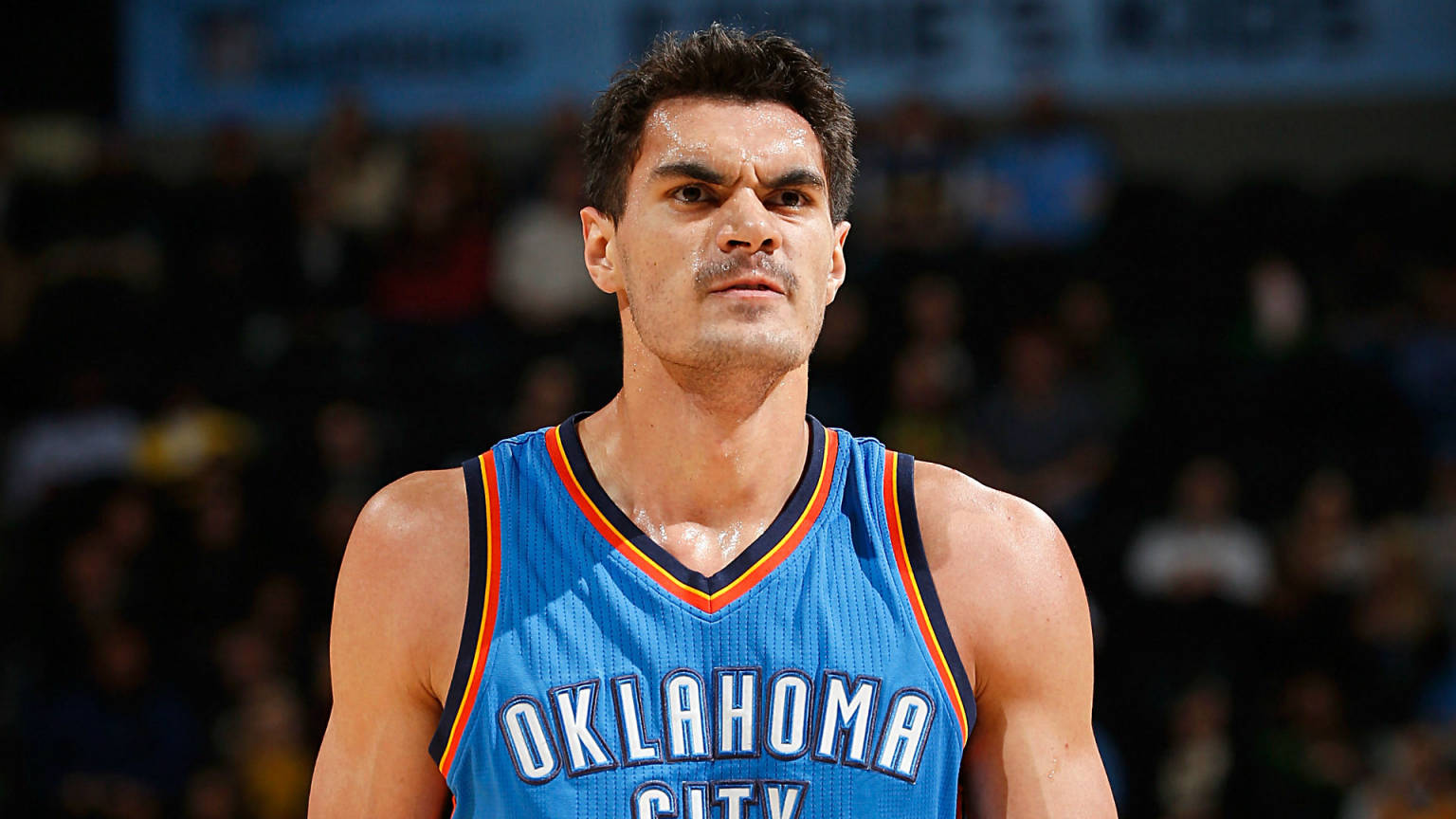 史蒂文·亞當斯 NBA OKC 壁紙 — 一名身穿藍色奧克拉荷馬城球衣的專注籃球運動員，在明亮的場館燈光下於比賽中凝視著。