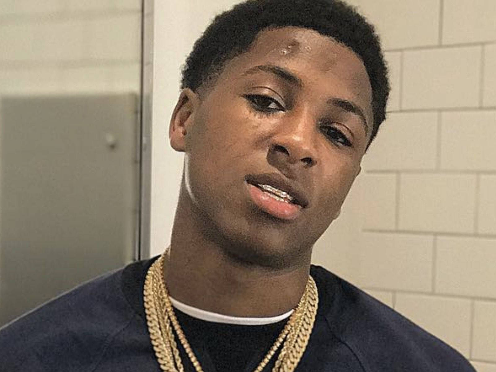 NBA YoungBoy — 自由之人 壁紙 — 一位短髮、戴金鏈的年輕男子在鋪滿瓷磚的房間裡直視鏡頭。