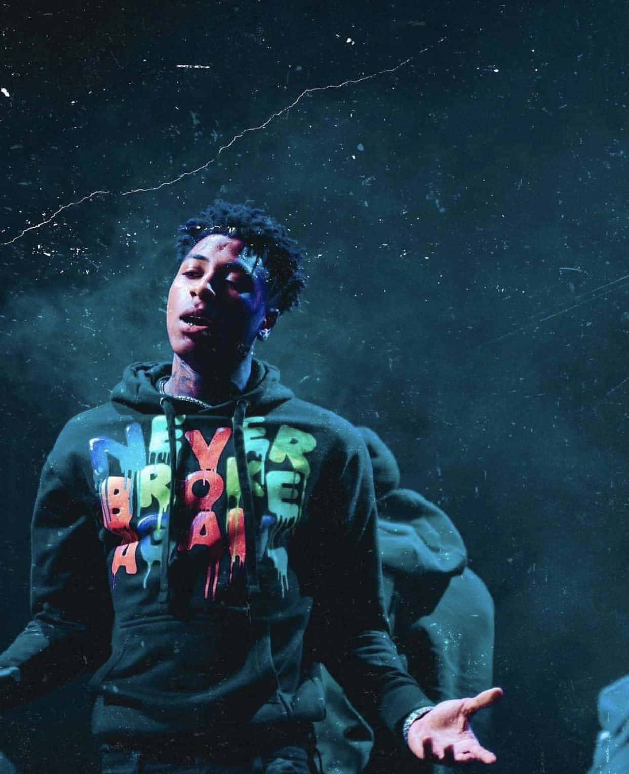 NBA Youngboy 「從未折損」主題頭像 壁紙 — 一個穿著深色連帽衫、帶有彩色滴落文字的年輕男子，站在煙霧繚繞的藍色背景前，燈光效果戲劇化。