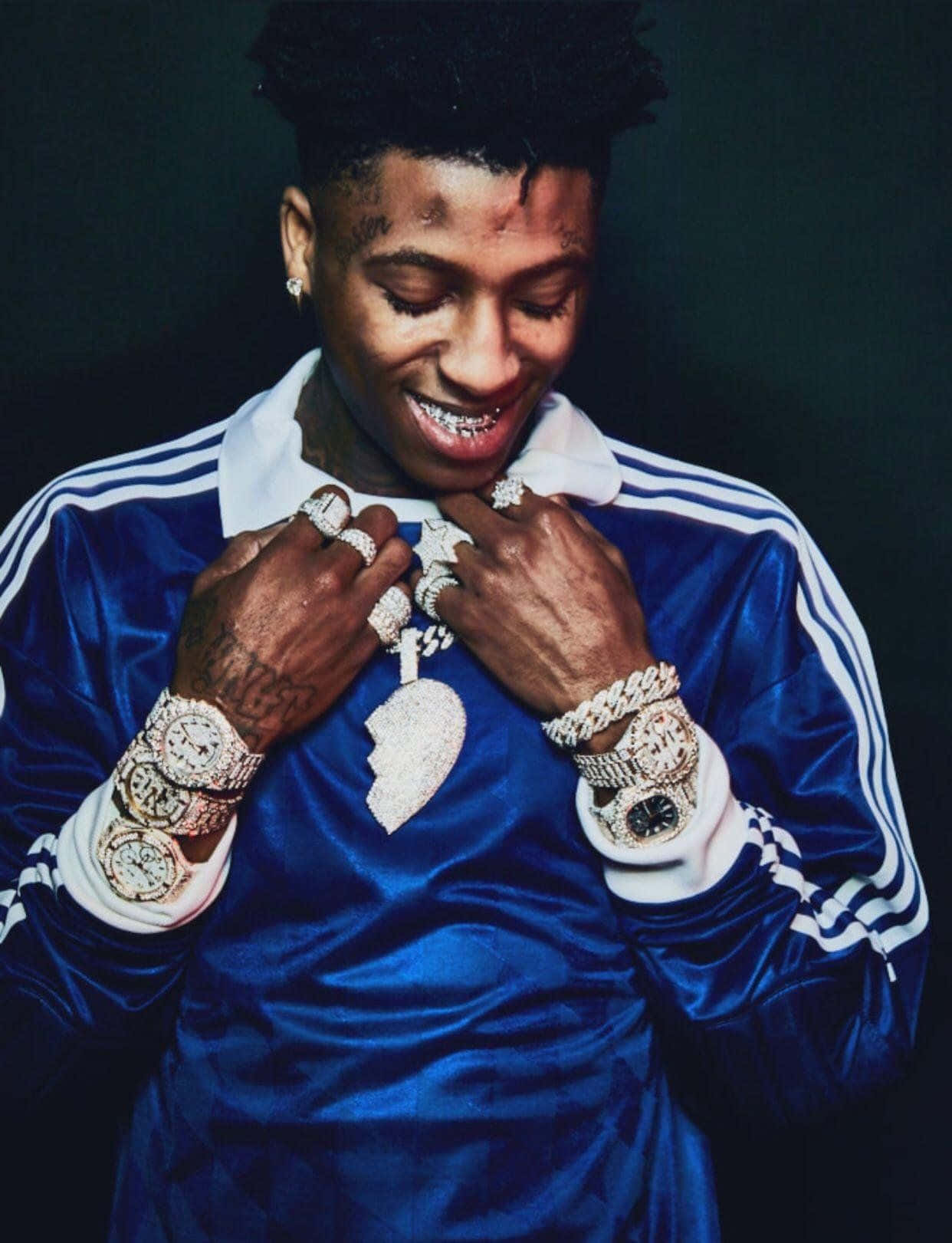 NBA Youngboy 閃耀珠寶頭像 壁紙 — 一名微笑的男子身穿藍色天鵝絨運動服，在深色背景前展示多枚鑲鑽手錶和戒指。