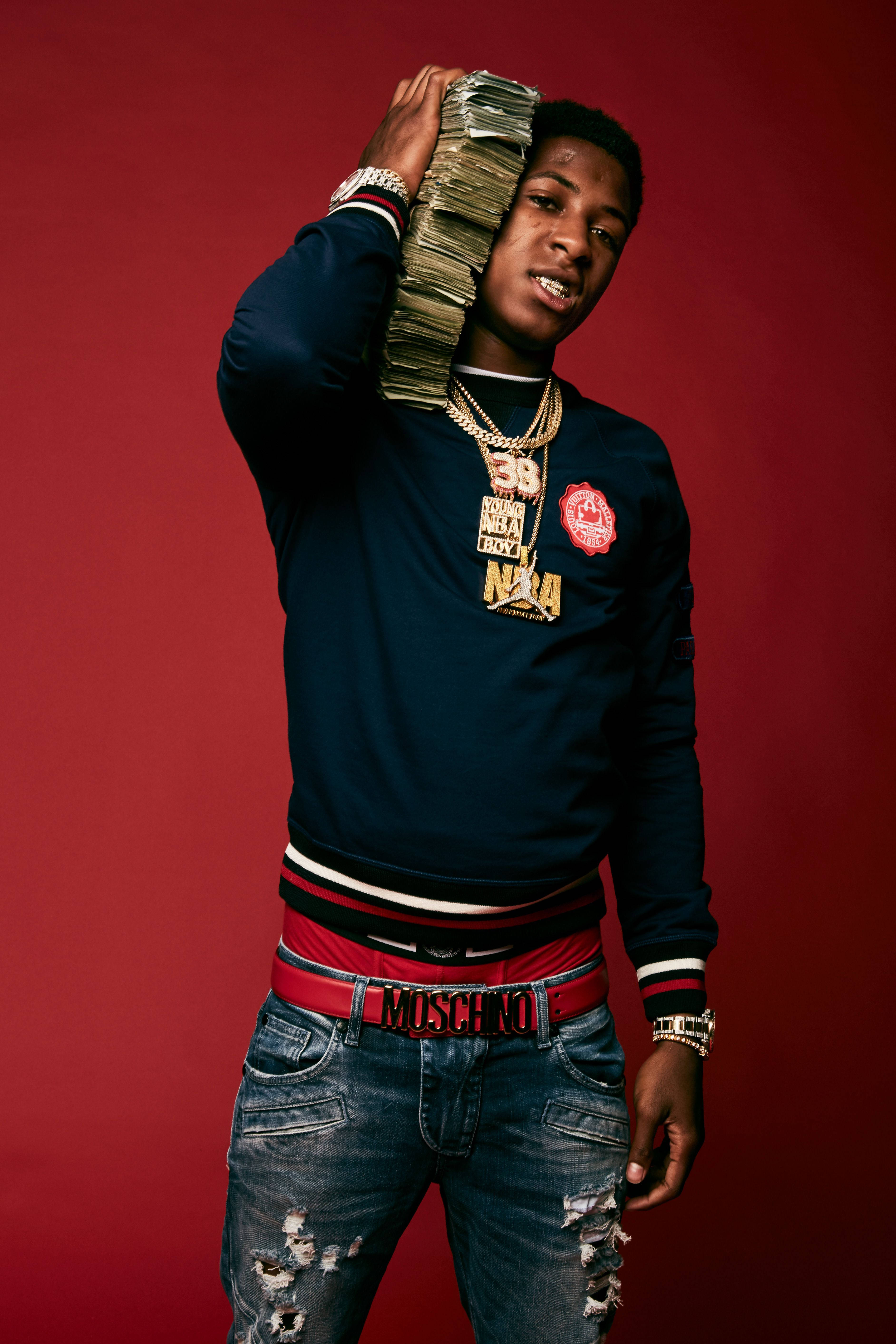 NBA Youngboy 身穿 Moschino 奢華服裝 壁紙 — 一名身穿海軍藍衛衣和破洞牛仔褲的年輕男子，在深紅色工作室背景前將一疊現金高舉過頭。