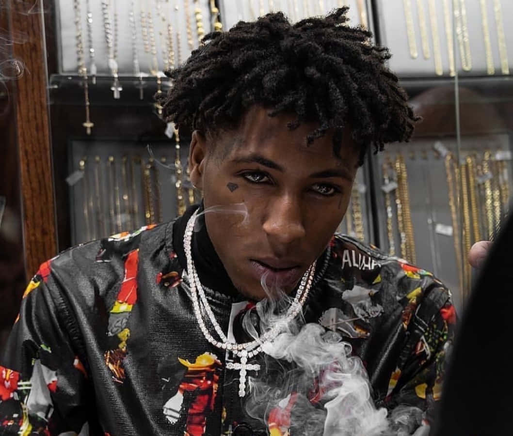 NBA Youngboy 在音樂與比賽之間 壁紙 — 一名年輕男子留著辮子，在珠寶店裡穿著色彩繽紛的夾克和鑽石鏈條，呼出煙霧。