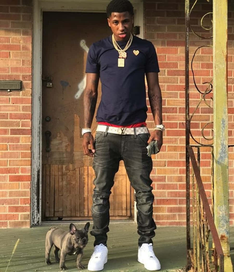 NBA Youngboy 奪取舞台 壁紙 — 一位身穿深藍色T恤和白色運動鞋的年輕男子自信地站在門廊上，身旁有一隻小灰色法國鬥牛犬。