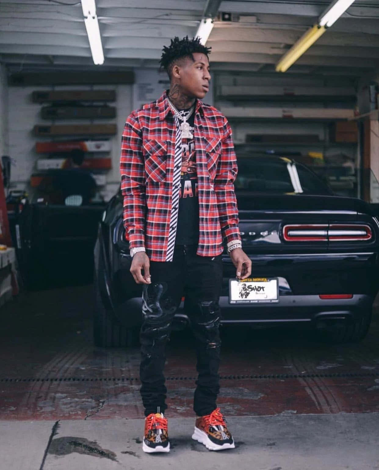 年輕男子穿著格仔襯衫和破洞牛仔褲，在車庫裡與黑色肌肉車合影。