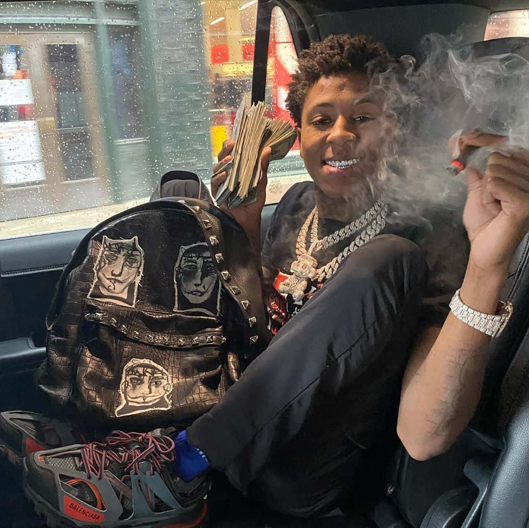 年輕男子在車內手持現金吸煙，帶有設計師背包和金鏈。 — 說唱歌手 壁紙