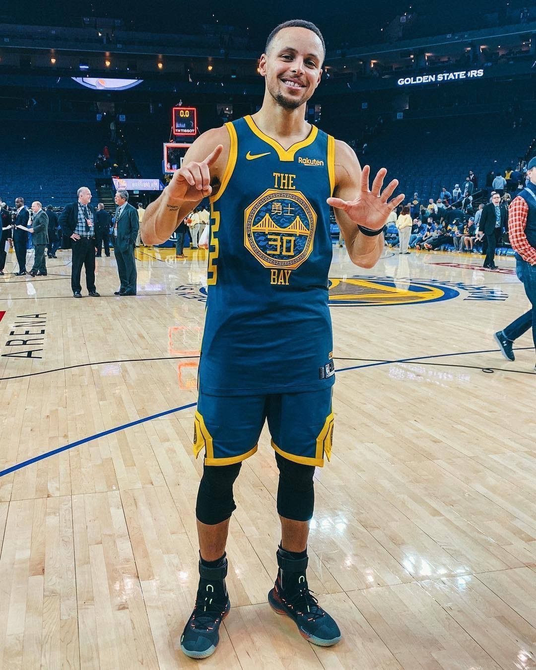 NBA 史蒂芬·柯瑞 iPhone 圖像 壁紙 — 籃球運動員史提芬居里身穿藍黃色金州勇士隊球衣，在拋光硬木球場上微笑並做手勢。