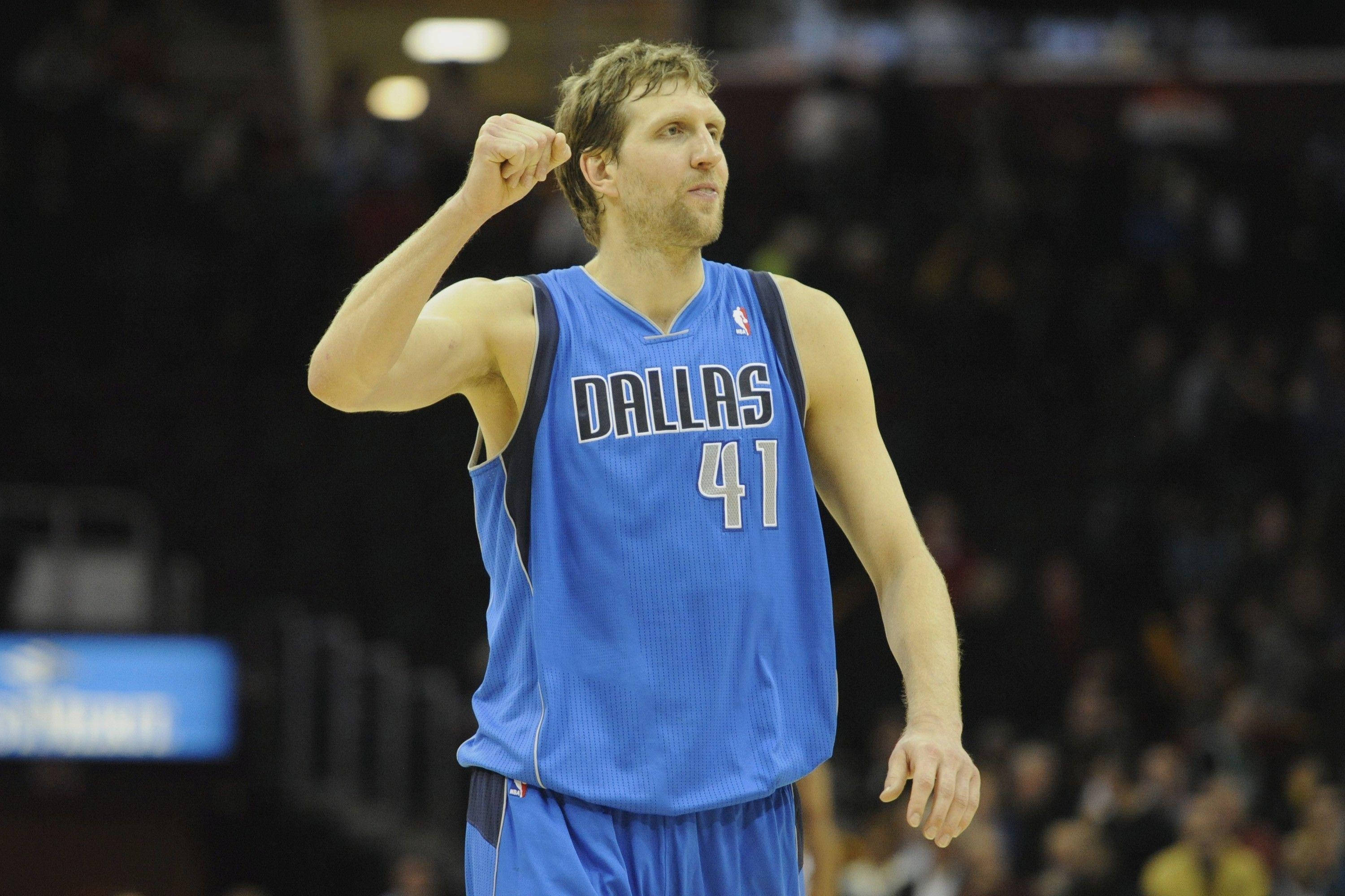 NBA 德克·諾維茨基 壁紙 — 一名身穿藍色達拉斯球衣的籃球運動員在場館燈光下的比賽中高舉拳頭慶祝勝利。