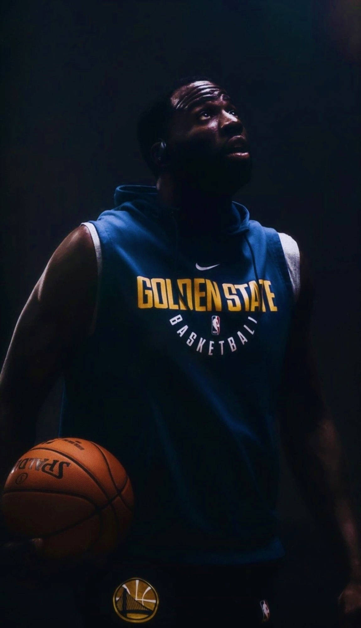 NBA 德雷蒙德·格林 壁紙 — 一名身穿金州勇士連帽衫的籃球運動員在黑暗背景下，於戲劇性的聚光燈下拿著一個Spalding籃球。