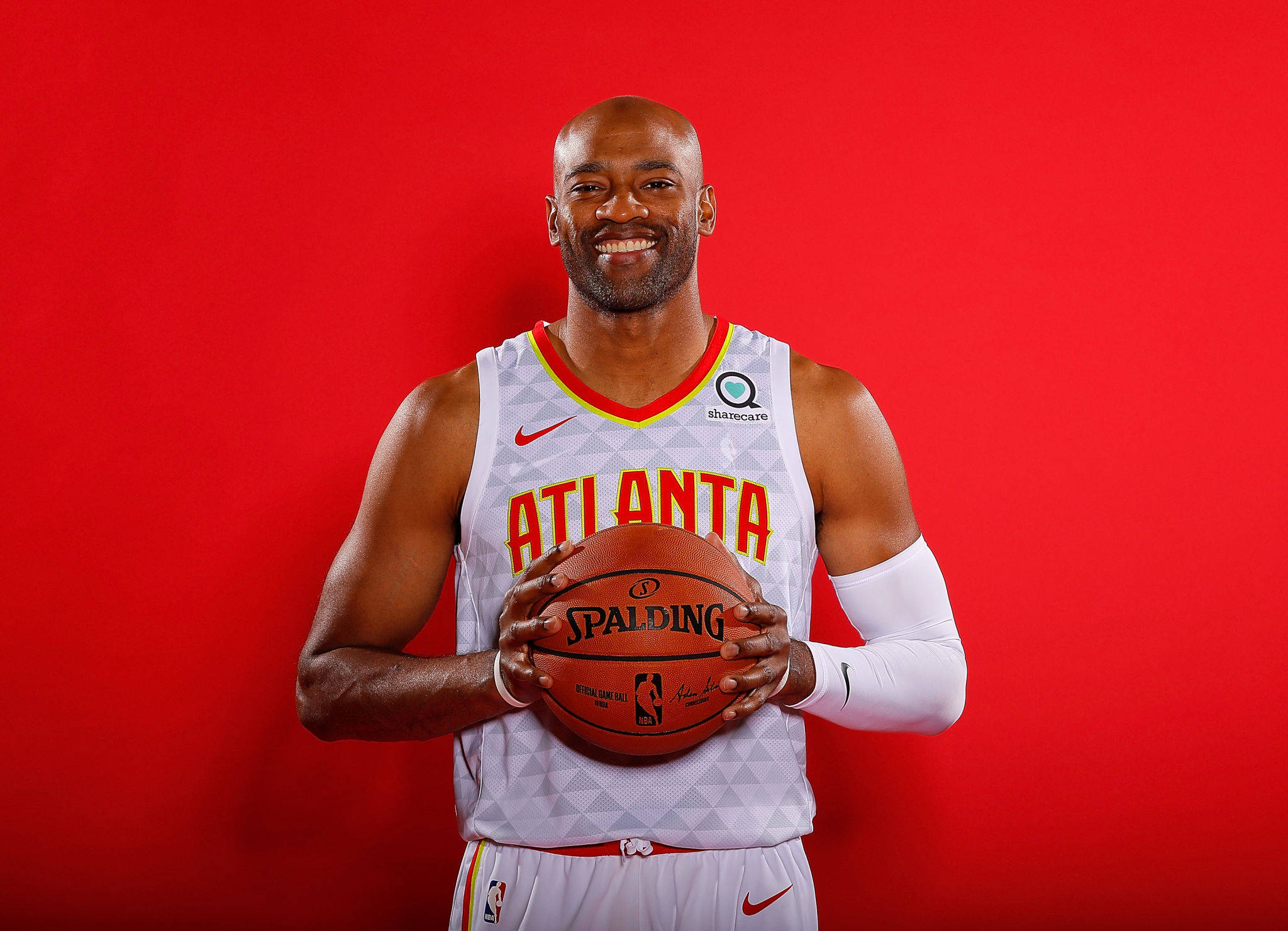 NBA 文斯·卡特 壁紙 — 一位微笑的籃球運動員身穿亞特蘭大球衣，在純紅色攝影棚背景前拿著一顆Spalding籃球。
