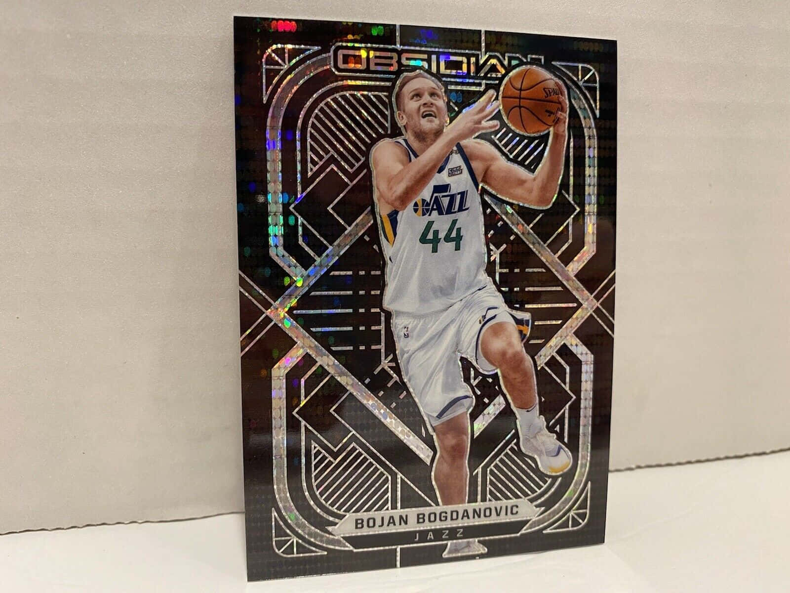 NBA 波揚·博格丹諾維奇 2021 年 Prizm 卡片 壁紙 — 博揚·博格達諾維奇身穿白色爵士隊球衣，在全息幾何背景下的籃球交易卡中景照。