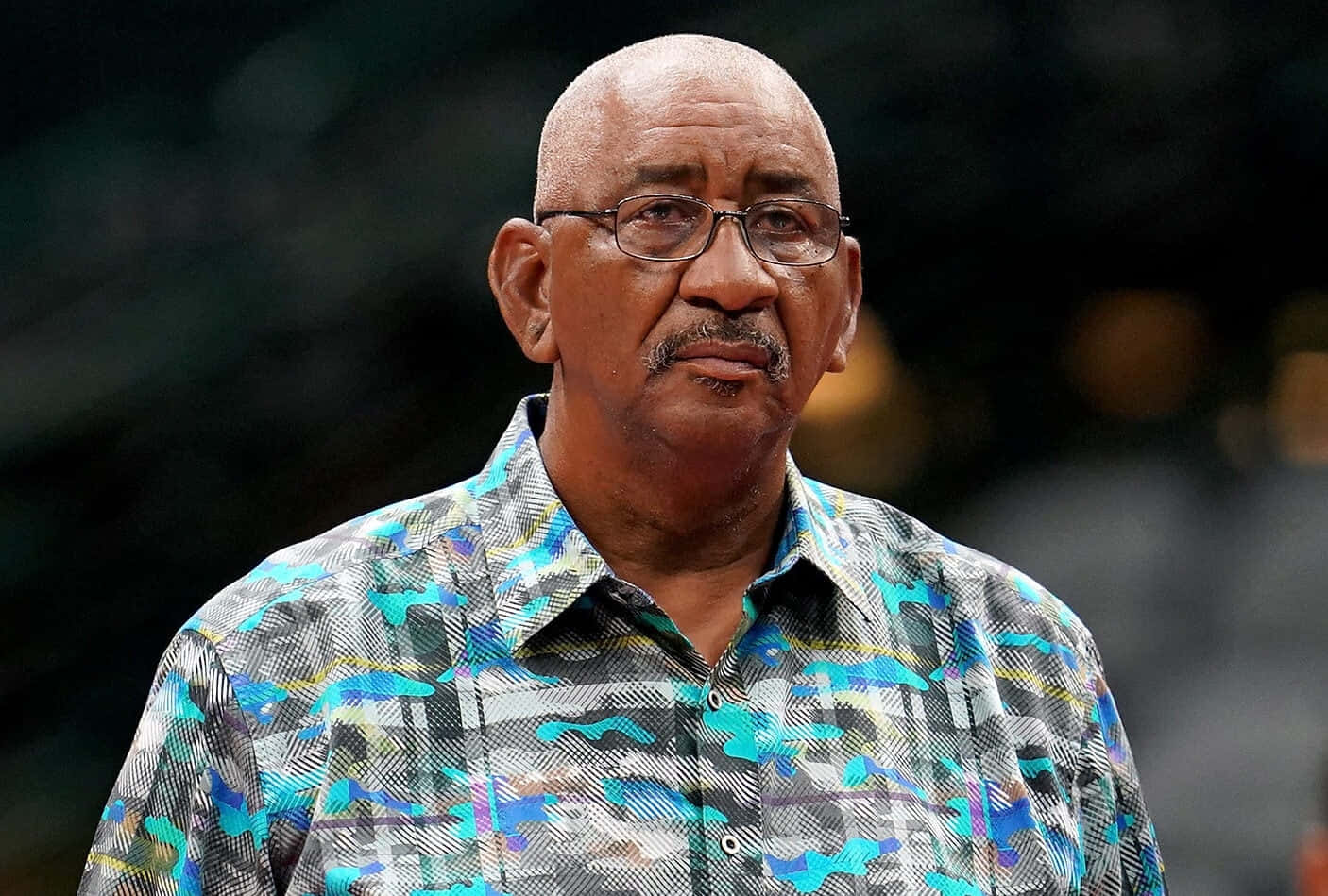 NBA傳奇喬治·格文籃球 壁紙 — 一位光頭戴眼鏡及鬍鬚的男子，穿著色彩繽紛的圖案襯衫，背景為模糊的室內環境。