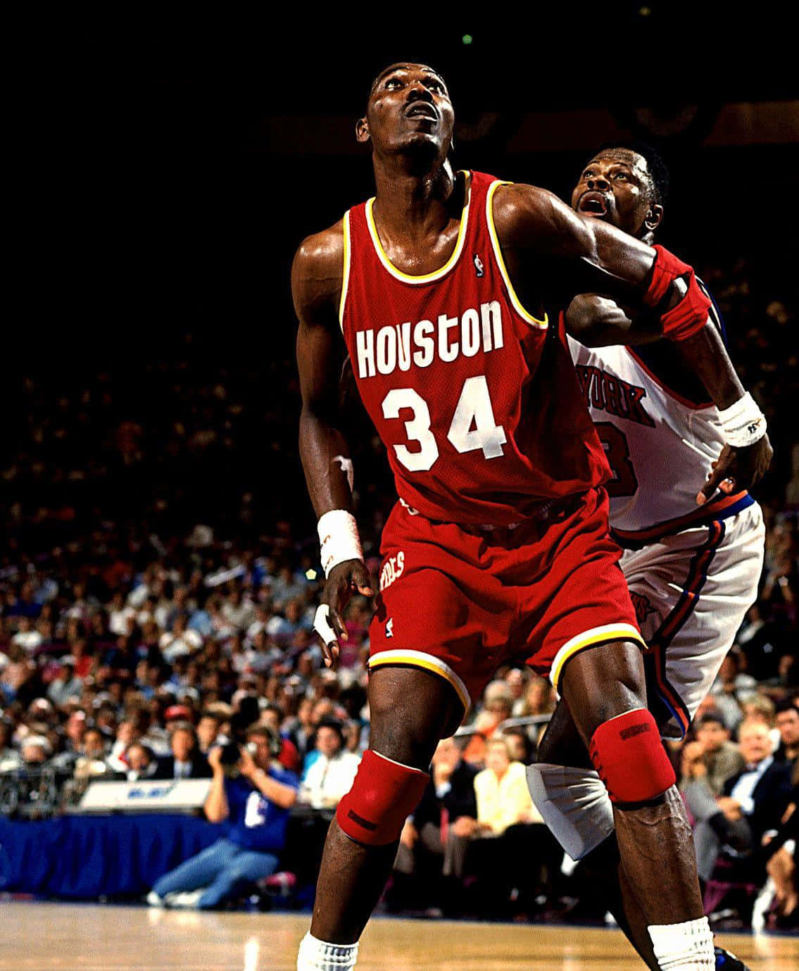 NBA傳奇對決：帕特里克·尤因對決哈基姆·奧拉朱旺 壁紙 — 一位身穿紅色34號球衣的侯斯頓火箭球員在擠滿觀眾的場館中跳起投籃，同時被防守。