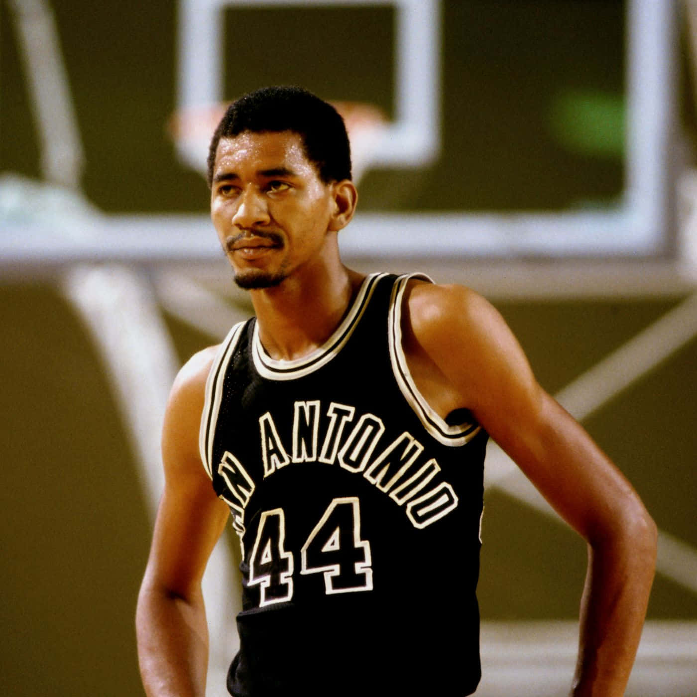 NBA傳奇球星喬治·格文籃球照 壁紙 — 一位專注的籃球運動員，身穿黑色聖安東尼奧球衣，背號44，站在球場上，背景的籃框模糊。