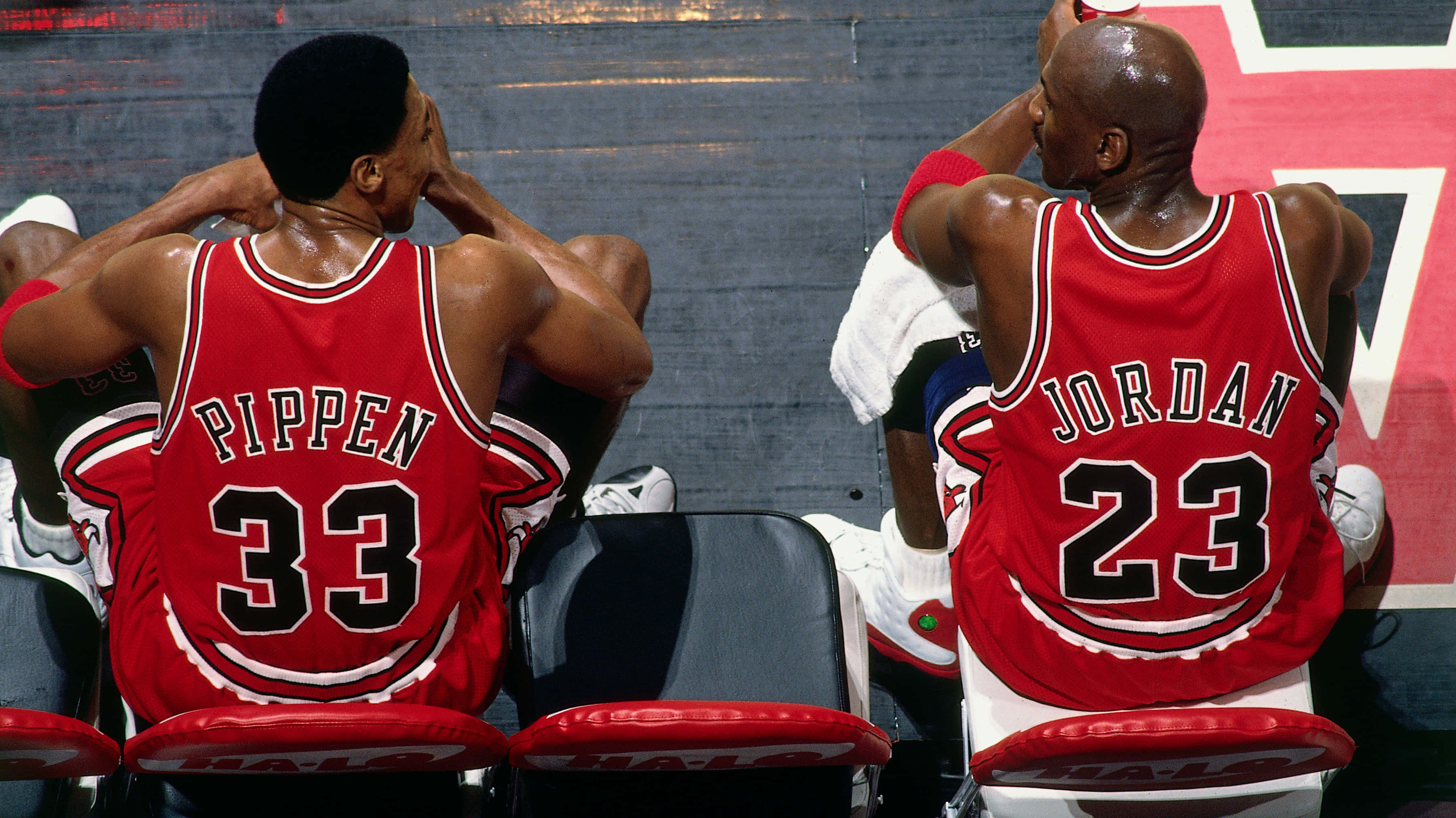 NBA傳奇邁克爾·喬丹 壁紙 — 兩名身穿紅色芝加哥公牛隊球衣的籃球運動員在比賽期間背對鏡頭坐在長凳上。