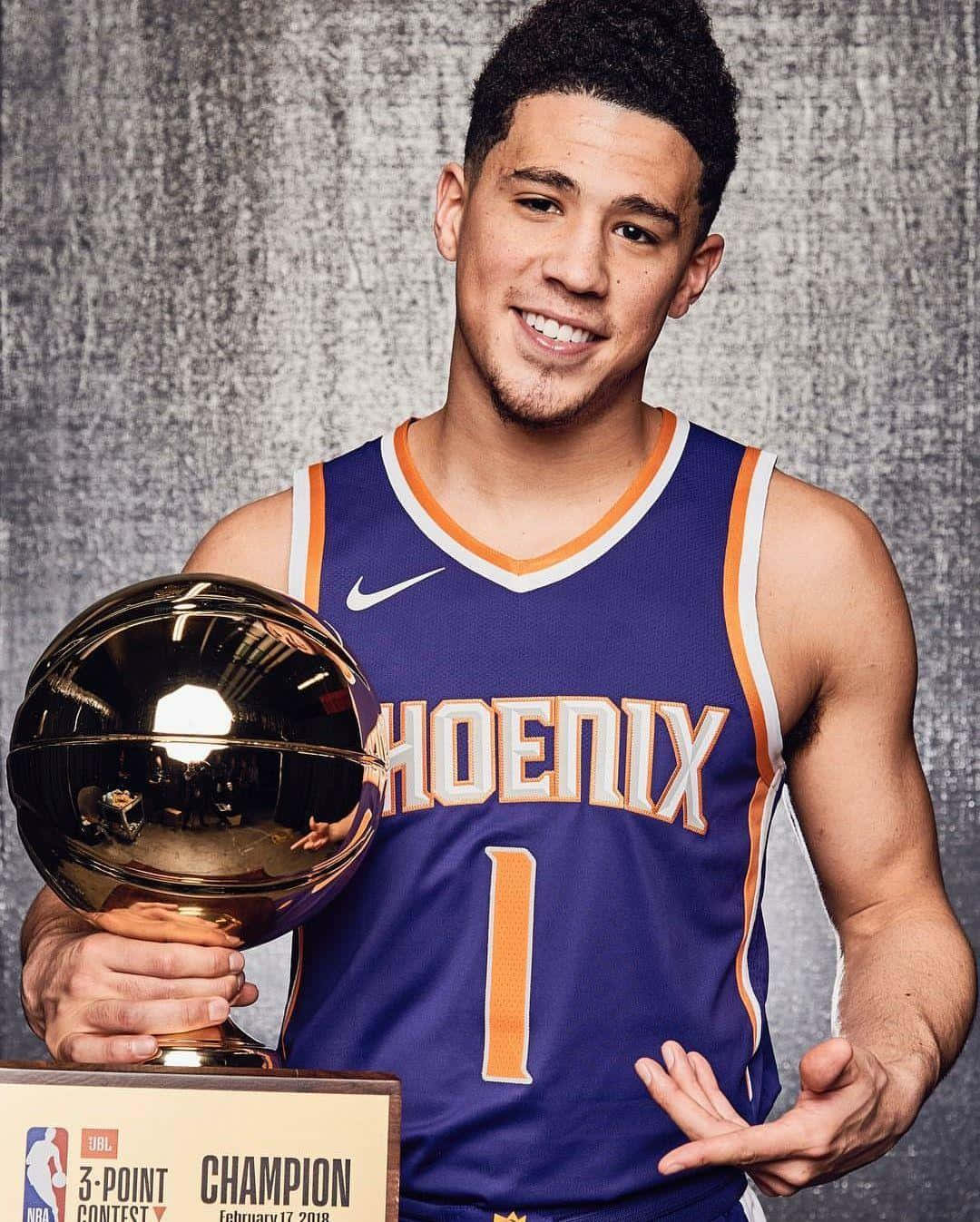 NBA全明星德文·布克與他的iPhone 壁紙 — 一位微笑的籃球運動員身穿鳳凰城太陽隊球衣，在紋理灰色背景前自豪地舉起閃閃發光的金色三分球大賽獎盃。